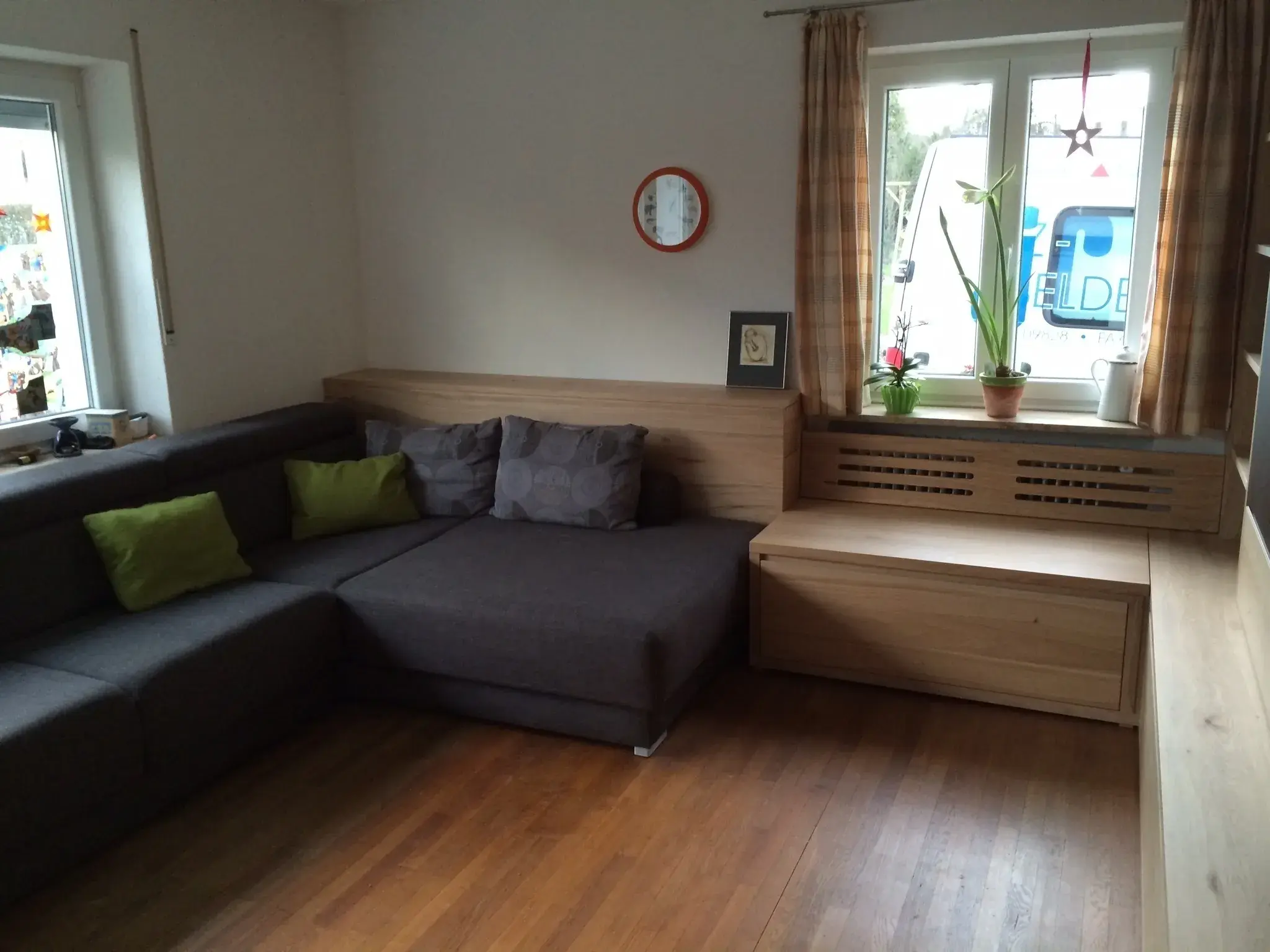 Wohnzimmer mit grauem Ecksofa, Holzboden und Fenster mit Pflanzen.