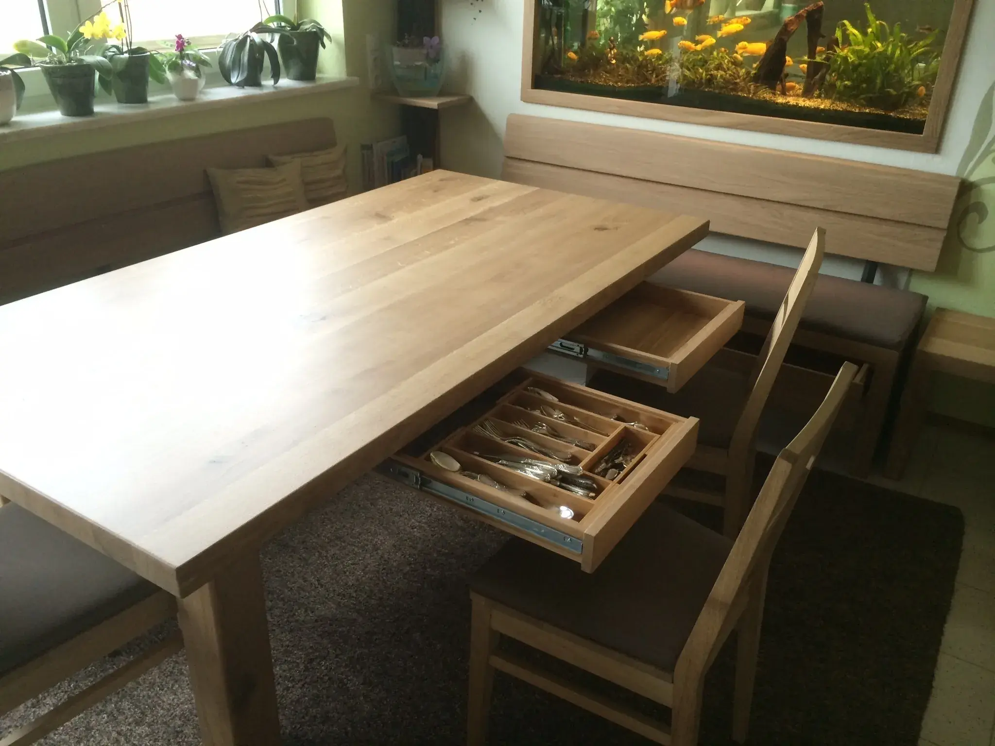 Holztisch mit geöffneten Schubladen voller Besteck, Stühle und Pflanzen am Fenster.