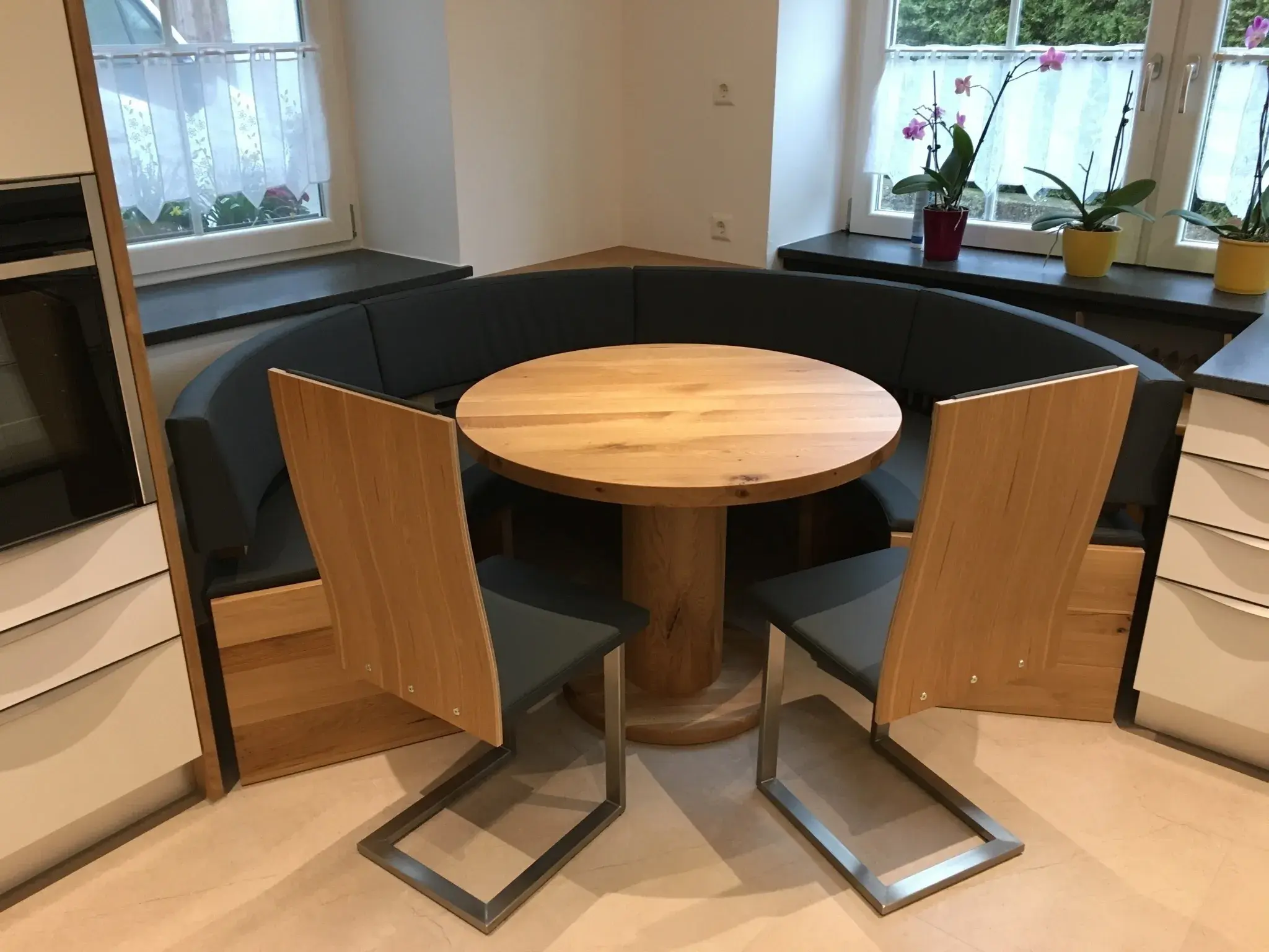 Runder Holztisch mit zwei Stühlen und einer Eckbank vor Fenstern mit Pflanzen.