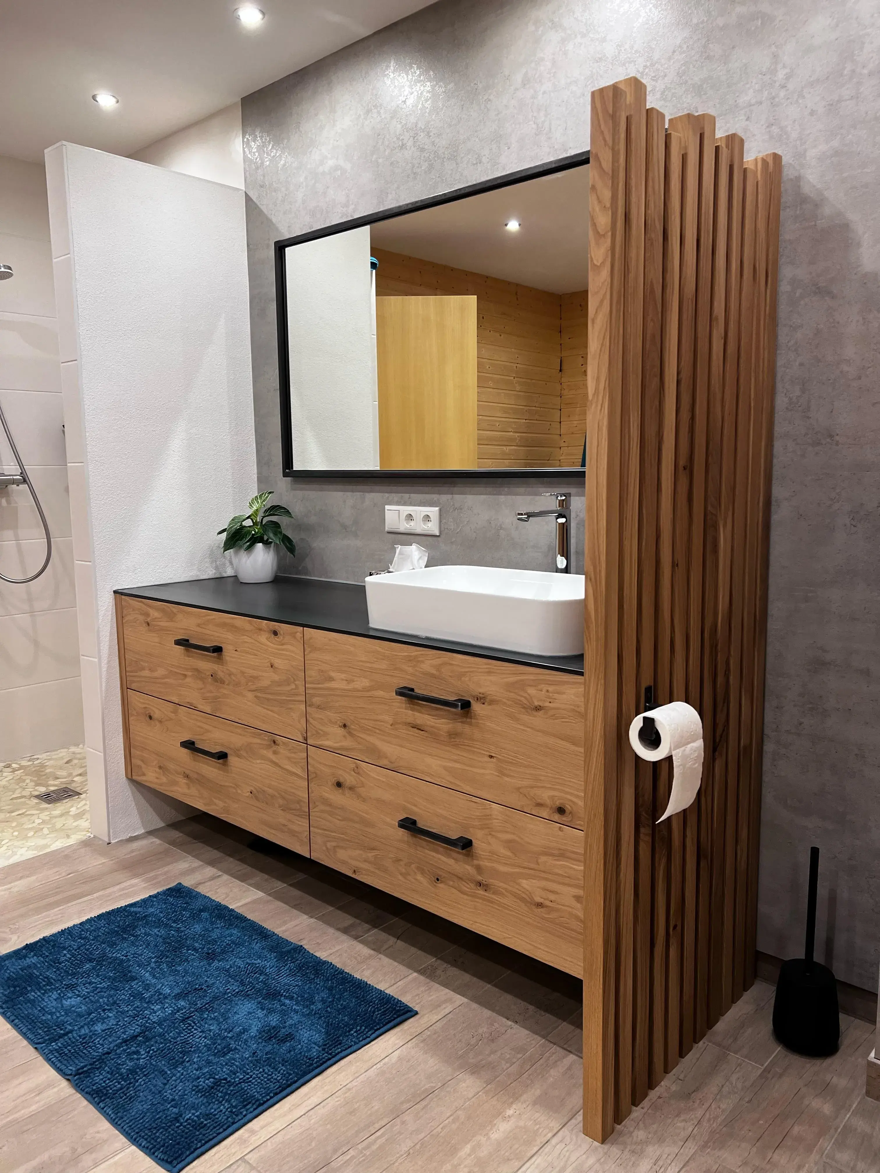 Modernes Badezimmer mit Holzmöbeln, großem Spiegel und blauem Teppich vor dem Waschbecken.