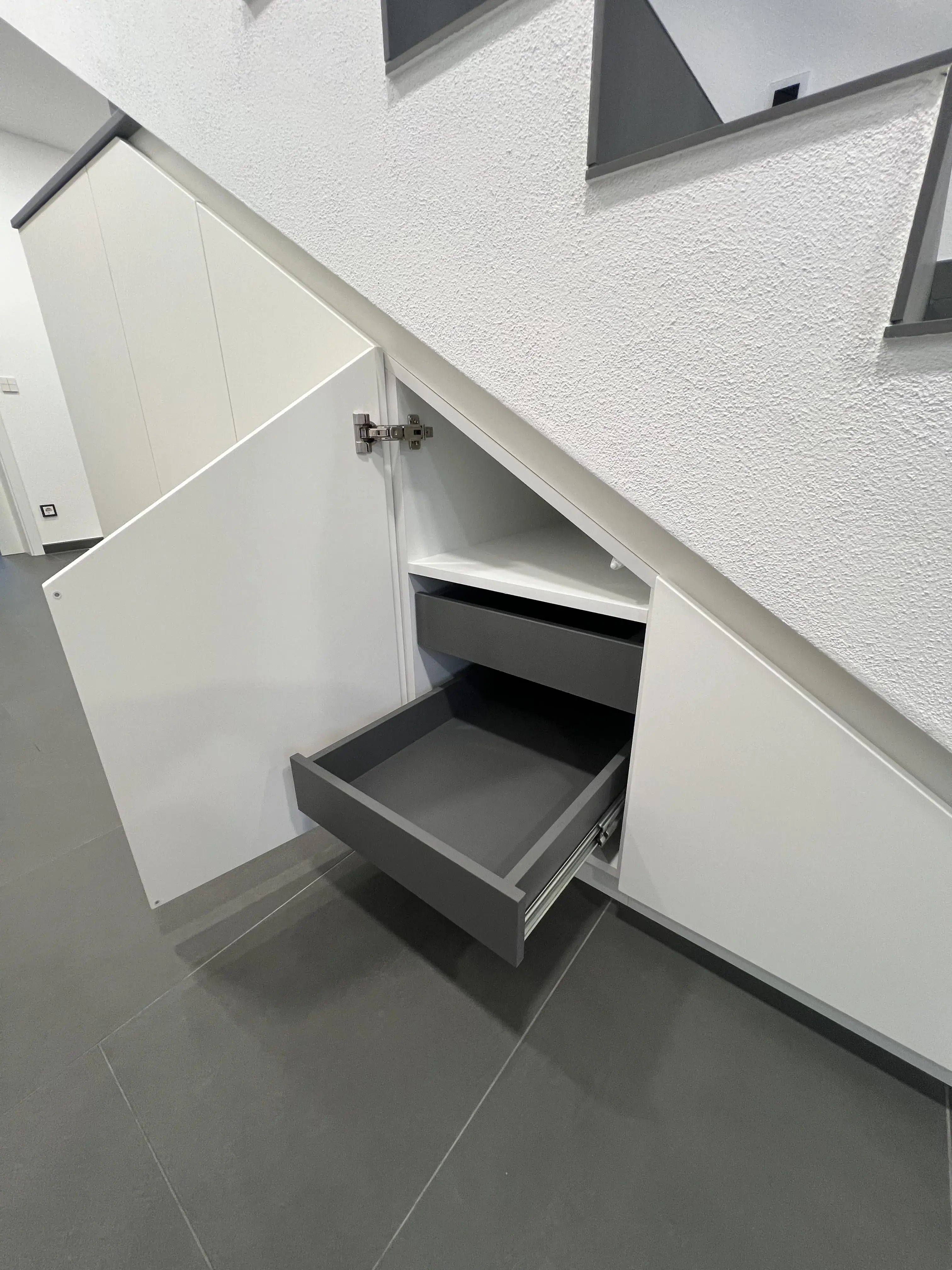 Graue Schubladen in weißem Schrank unter einer Treppe, eine ist geöffnet.