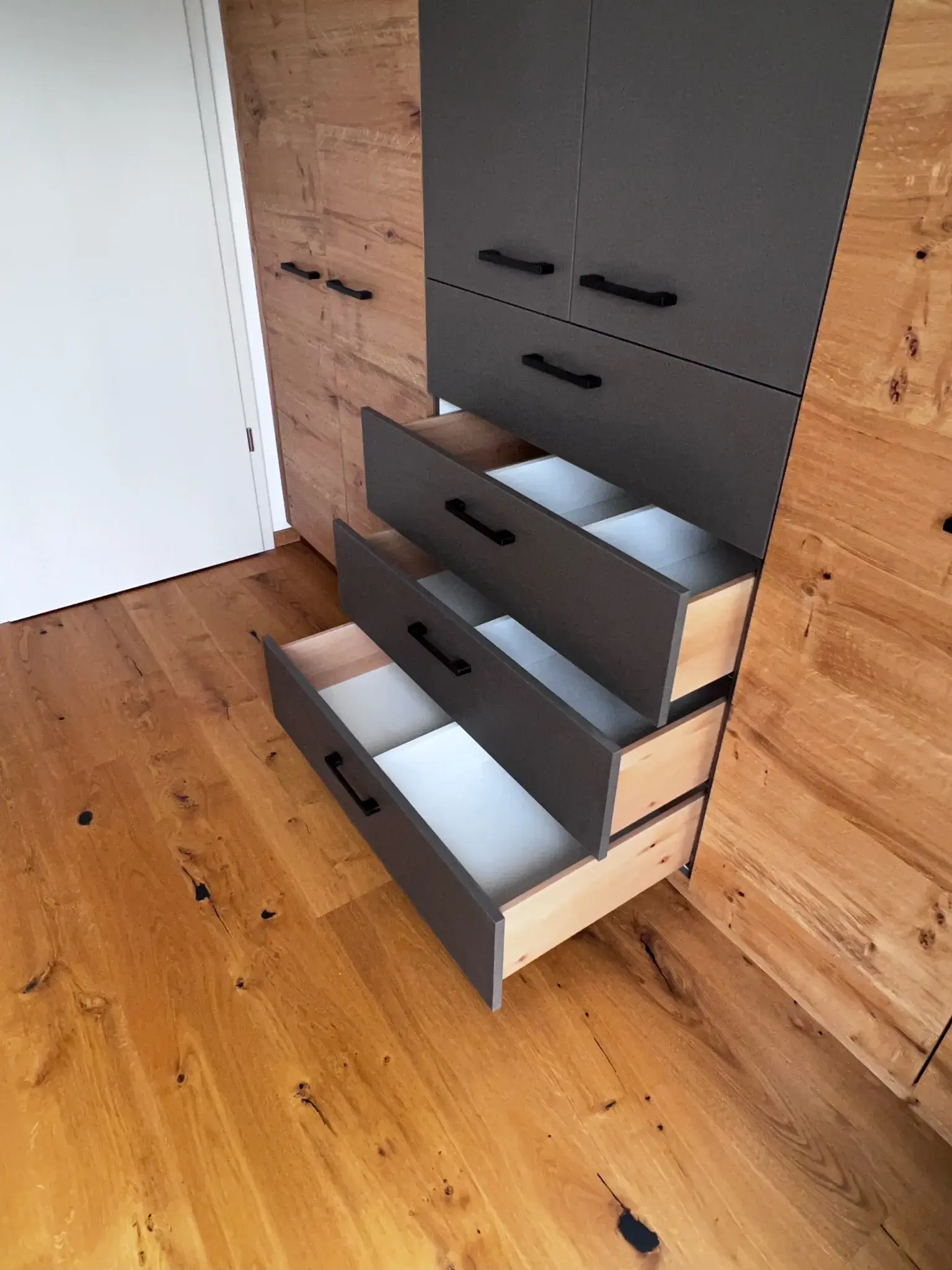 Vier geöffnete Schubladen in einem grauen Holzschrank auf Parkettboden.