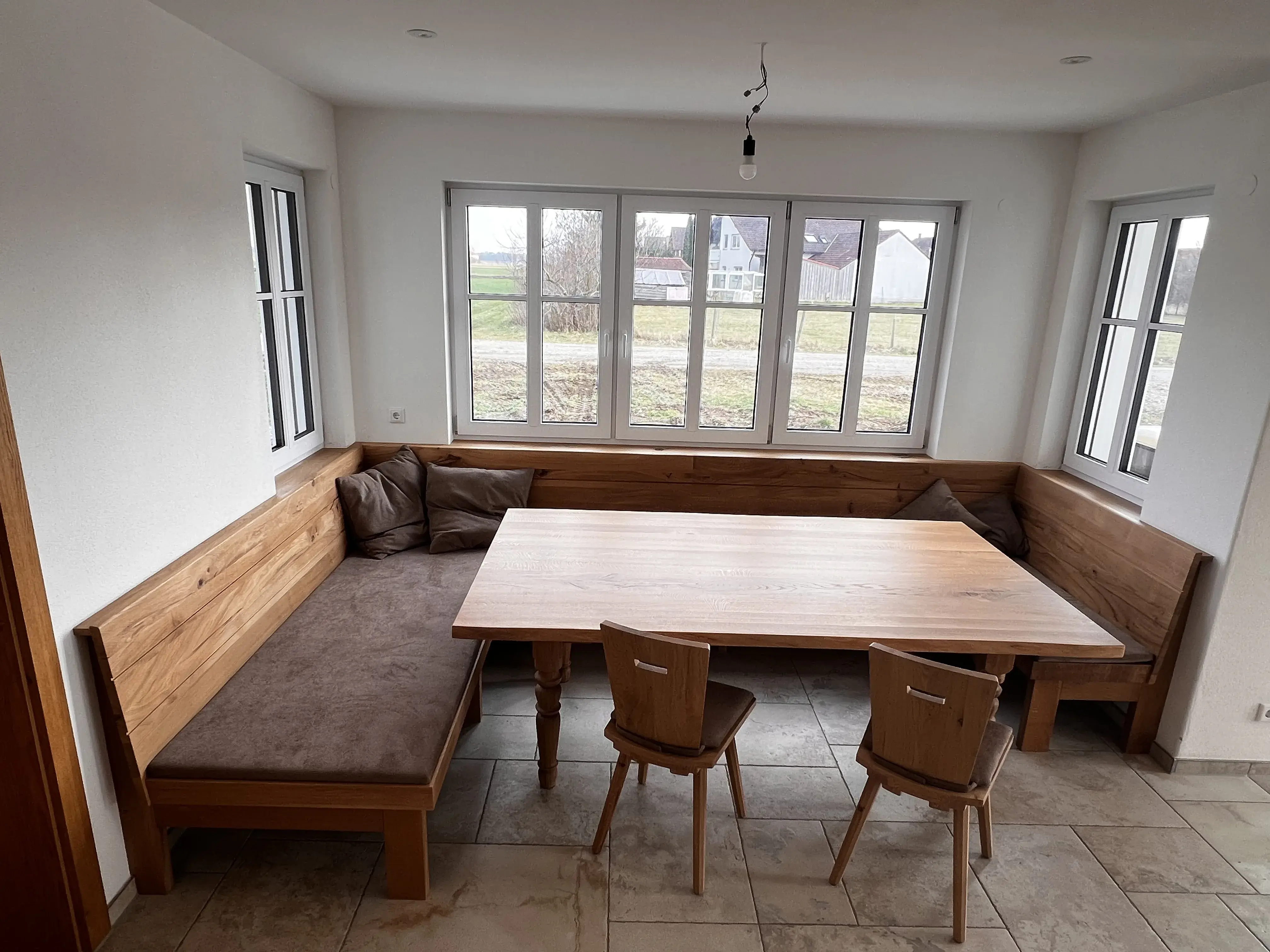 Ecke mit Holztisch, Eckbank und zwei Stühlen, helle Fenster im Hintergrund.