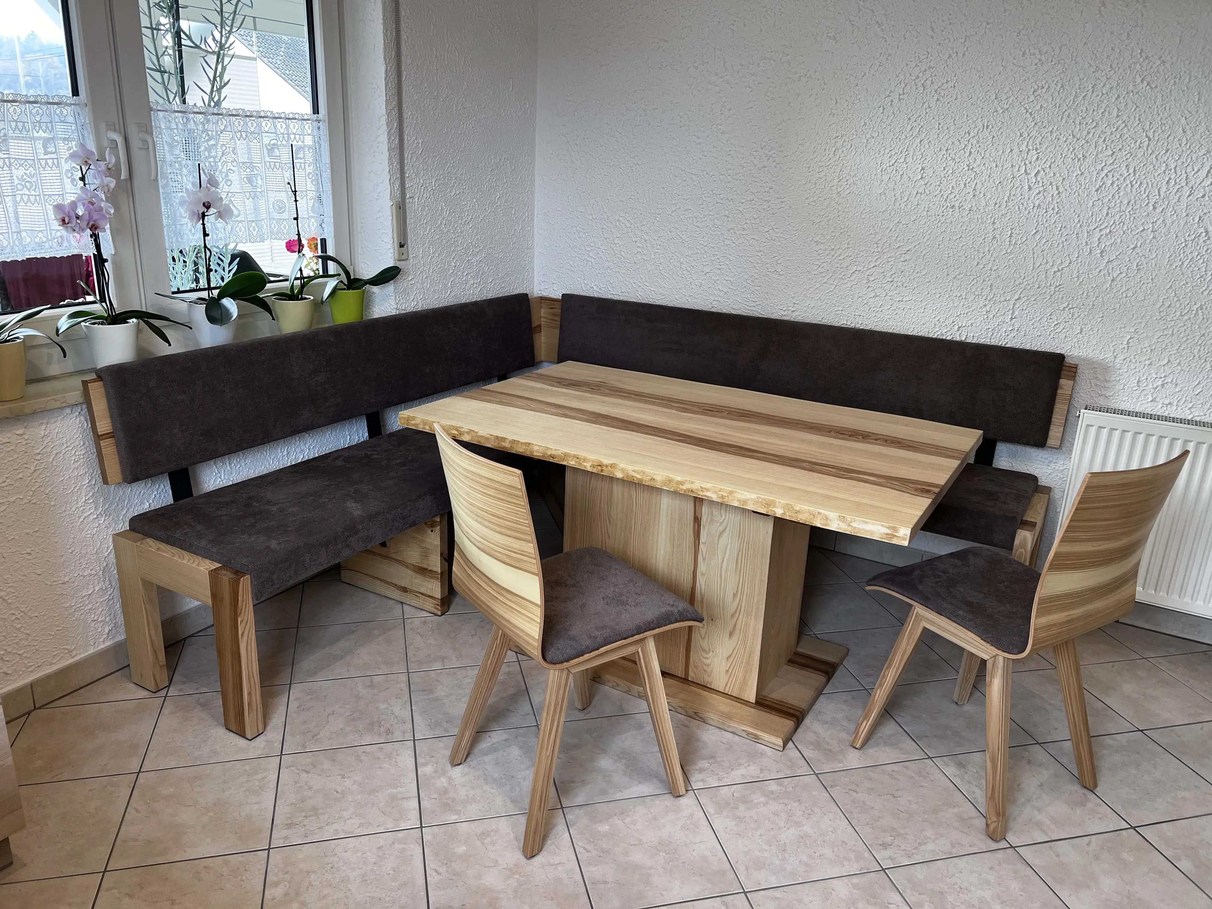 Eckbankgruppe aus Holz mit braunen Polstern, zwei Stühlen und Esstisch in heller Küche.