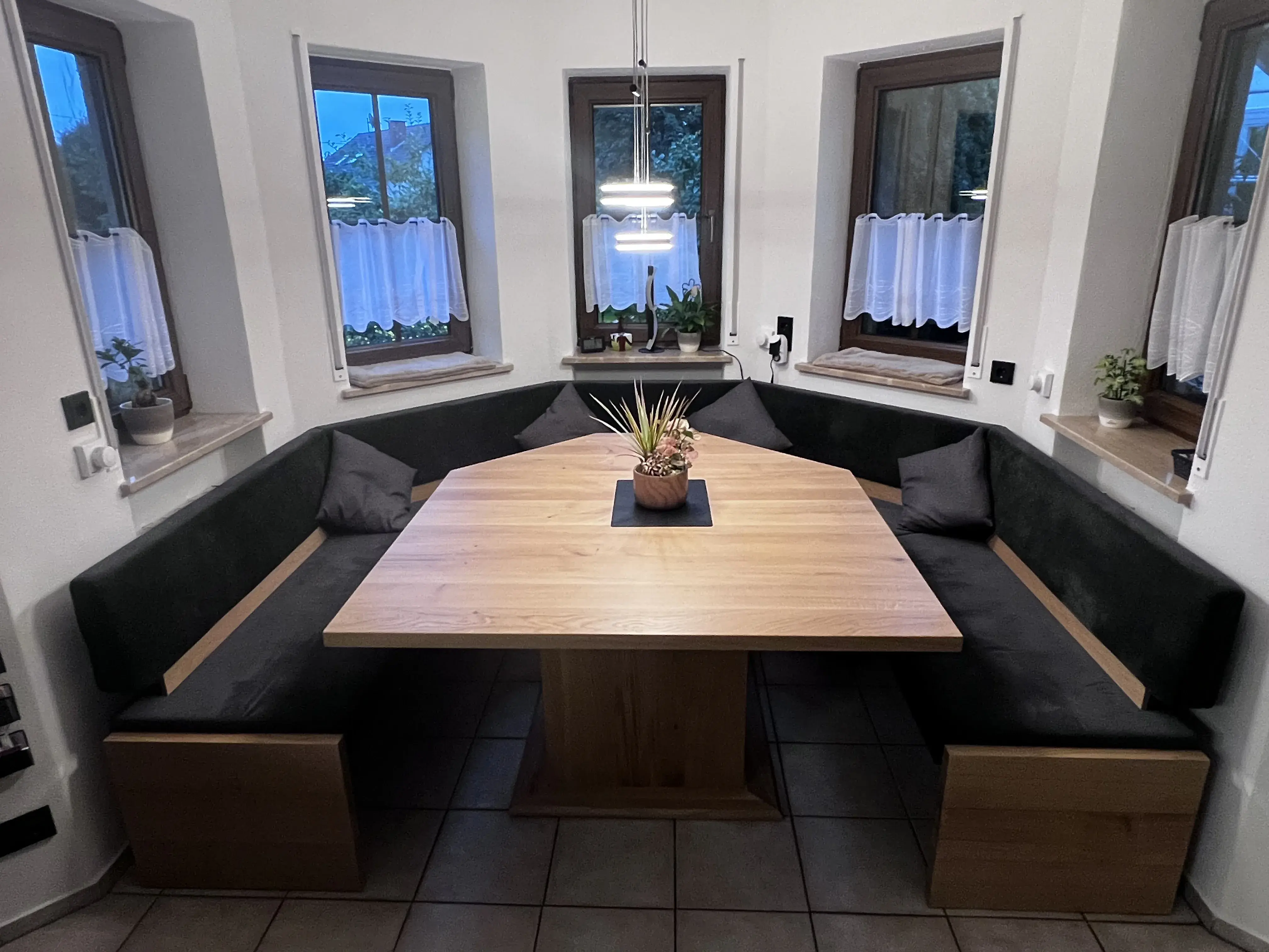 Eckbank aus Holz mit großem Tisch, Pflanze in der Mitte und drei Fenstern im Hintergrund.