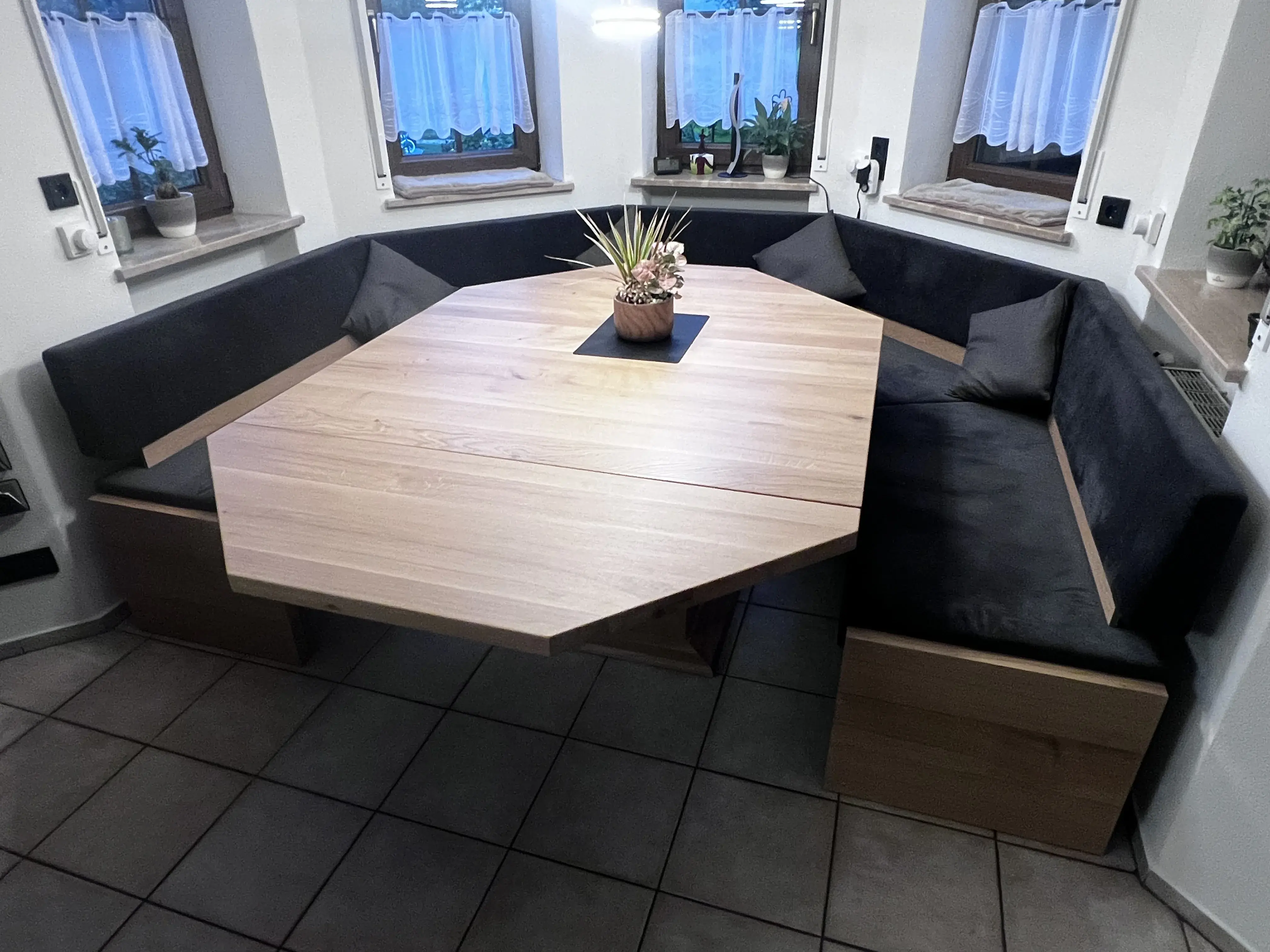 Eckbank mit achteckigem Holztisch und Blumentopf, umgeben von Fenstern mit Gardinen.