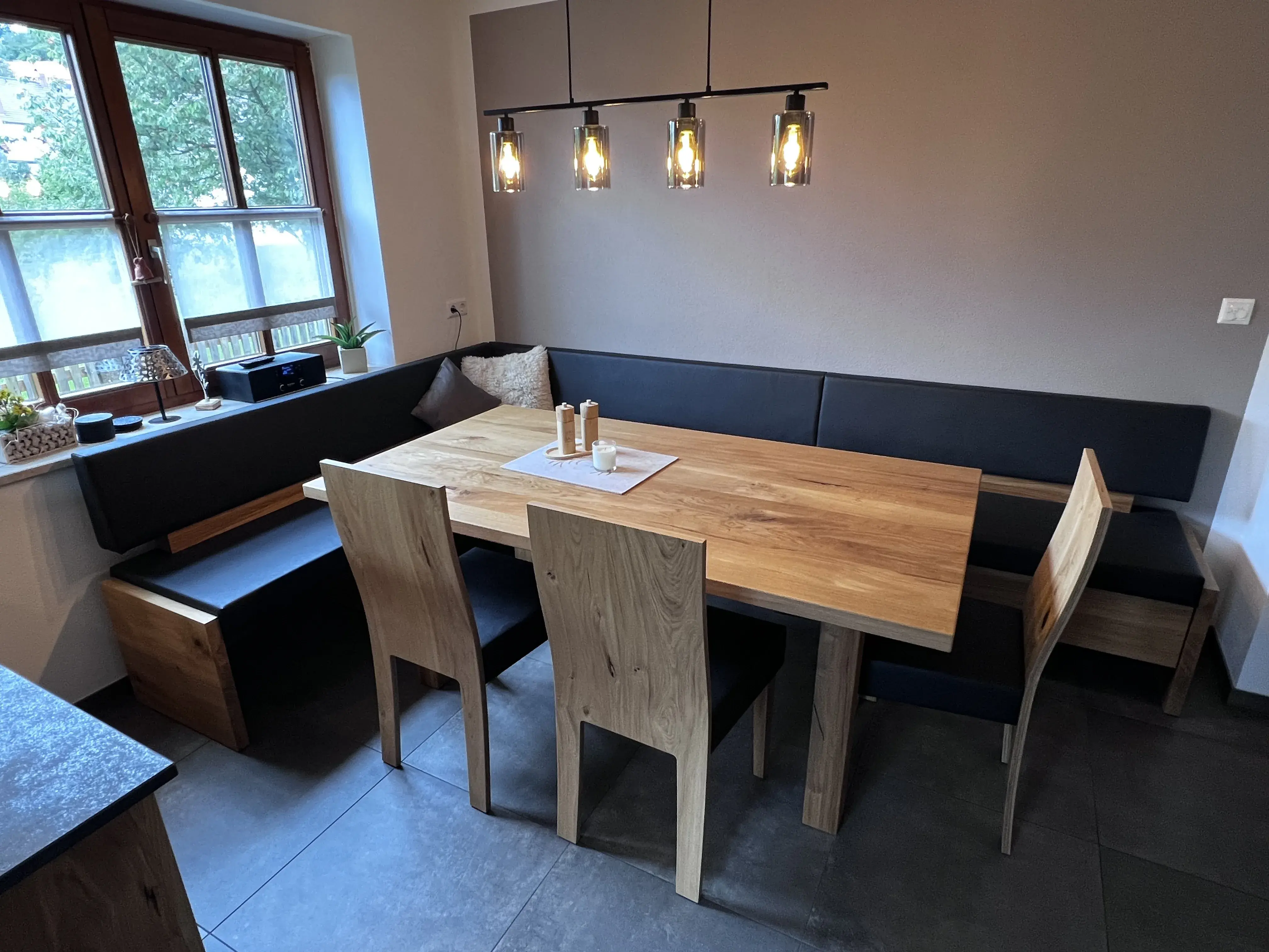 Holztisch mit Eckbank und Stühlen in modernem Esszimmer, Lampen darüber, Fenster links.