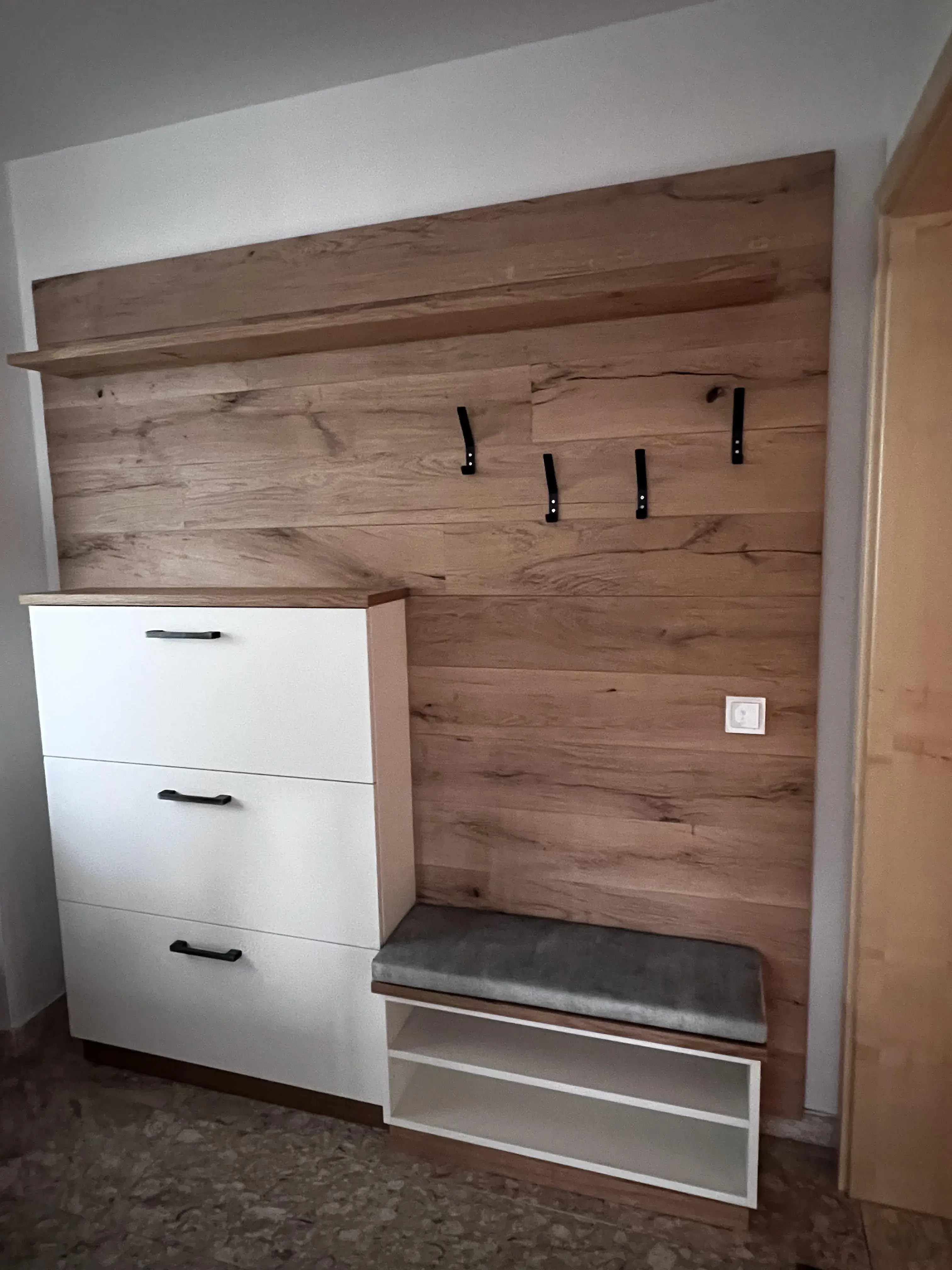 Garderobe mit Holzwand, Haken, Bank mit grauem Polster und weißem Schuhschrank.