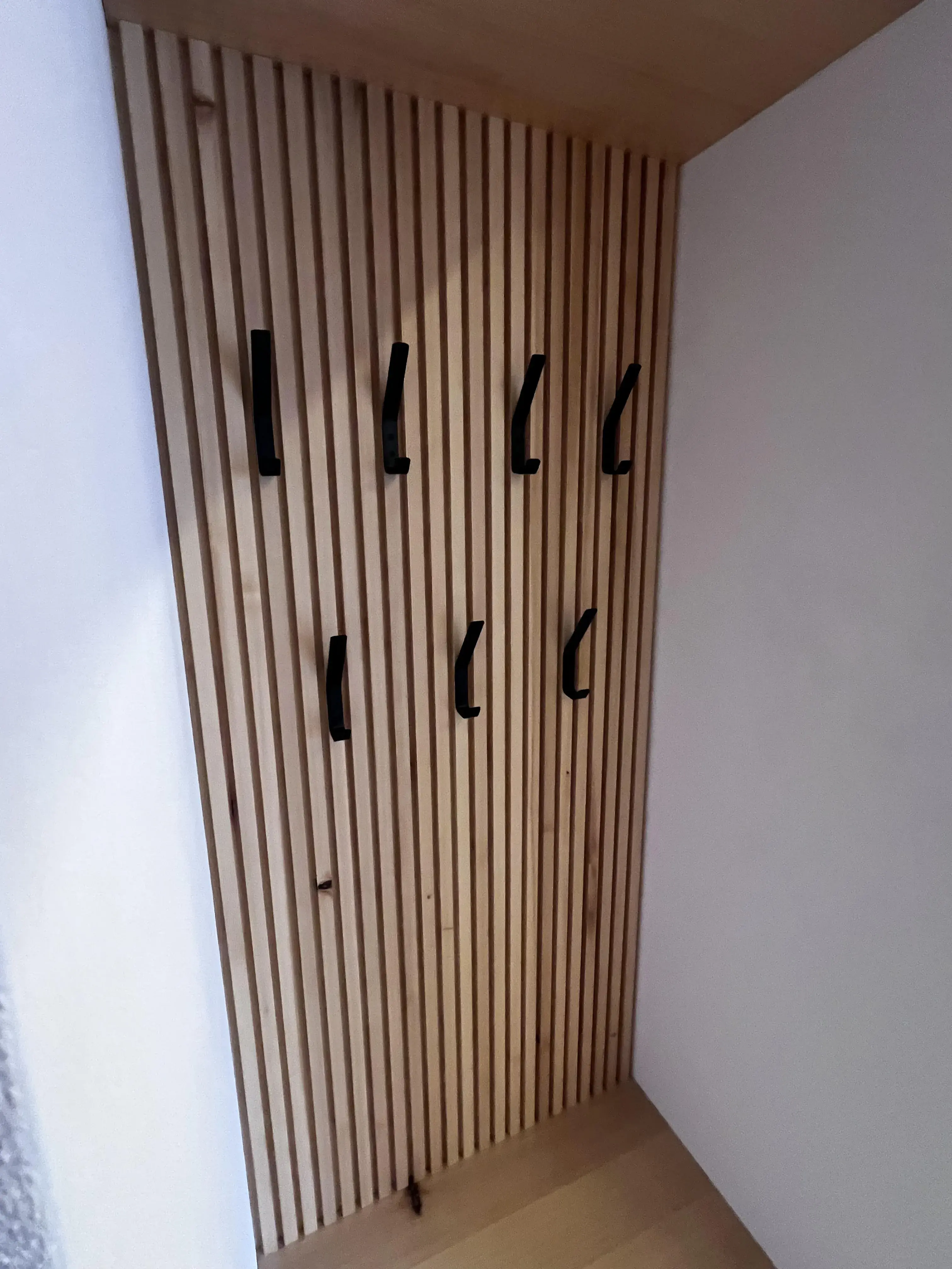 Holzwand mit sechs schwarzen Kleiderhaken in zwei Reihen.