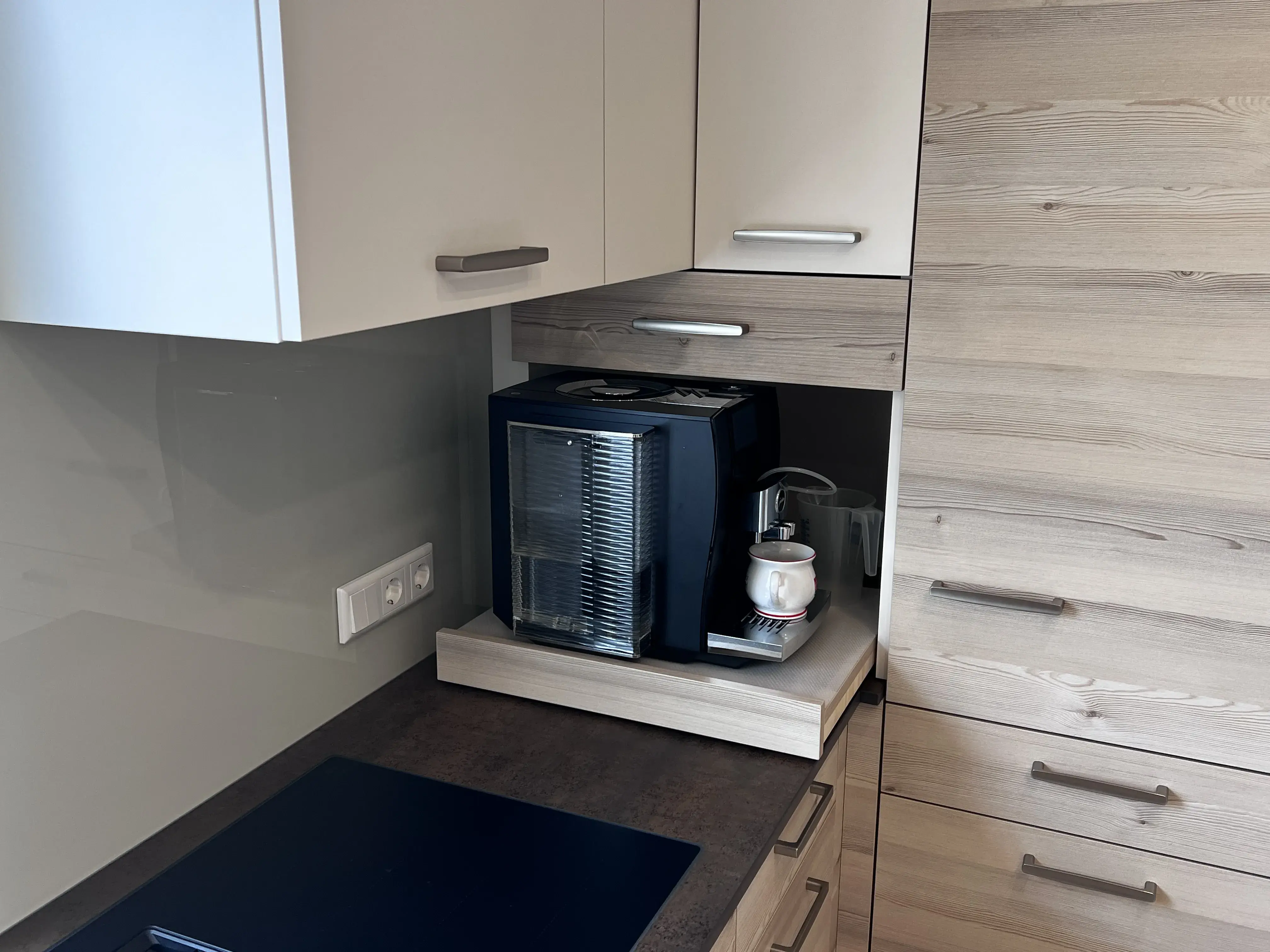 Kaffeemaschine auf ausziehbarem Regal in moderner Küche, daneben Tassen und Milchaufschäumer.