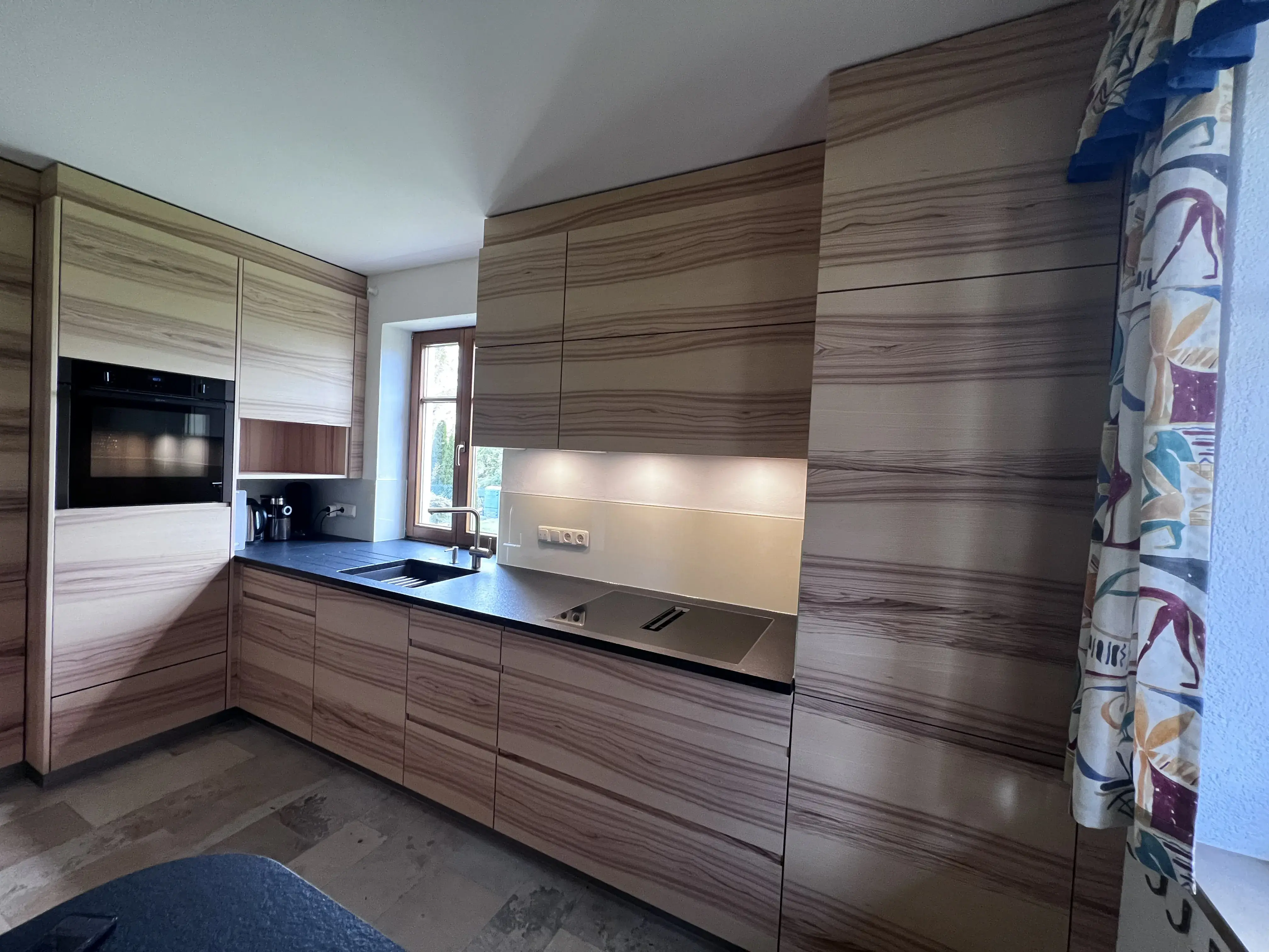 Moderne Holzküche mit schwarzen Arbeitsflächen und eingebauten Geräten, Tageslicht durch Fenster.