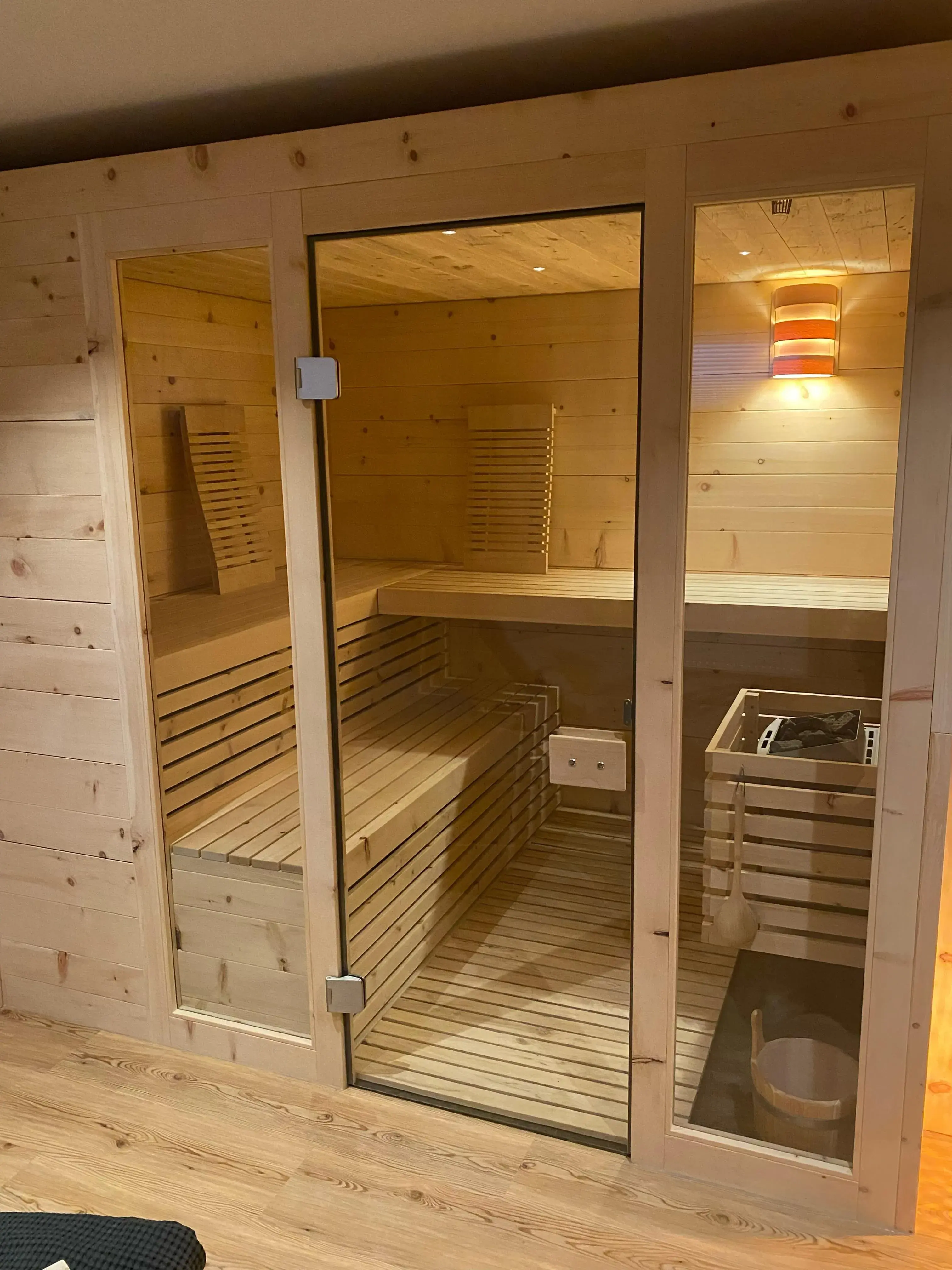 Holzsauna mit Glasfront, Innenraum beleuchtet, Holzbänke und Aufgusseimer sichtbar.