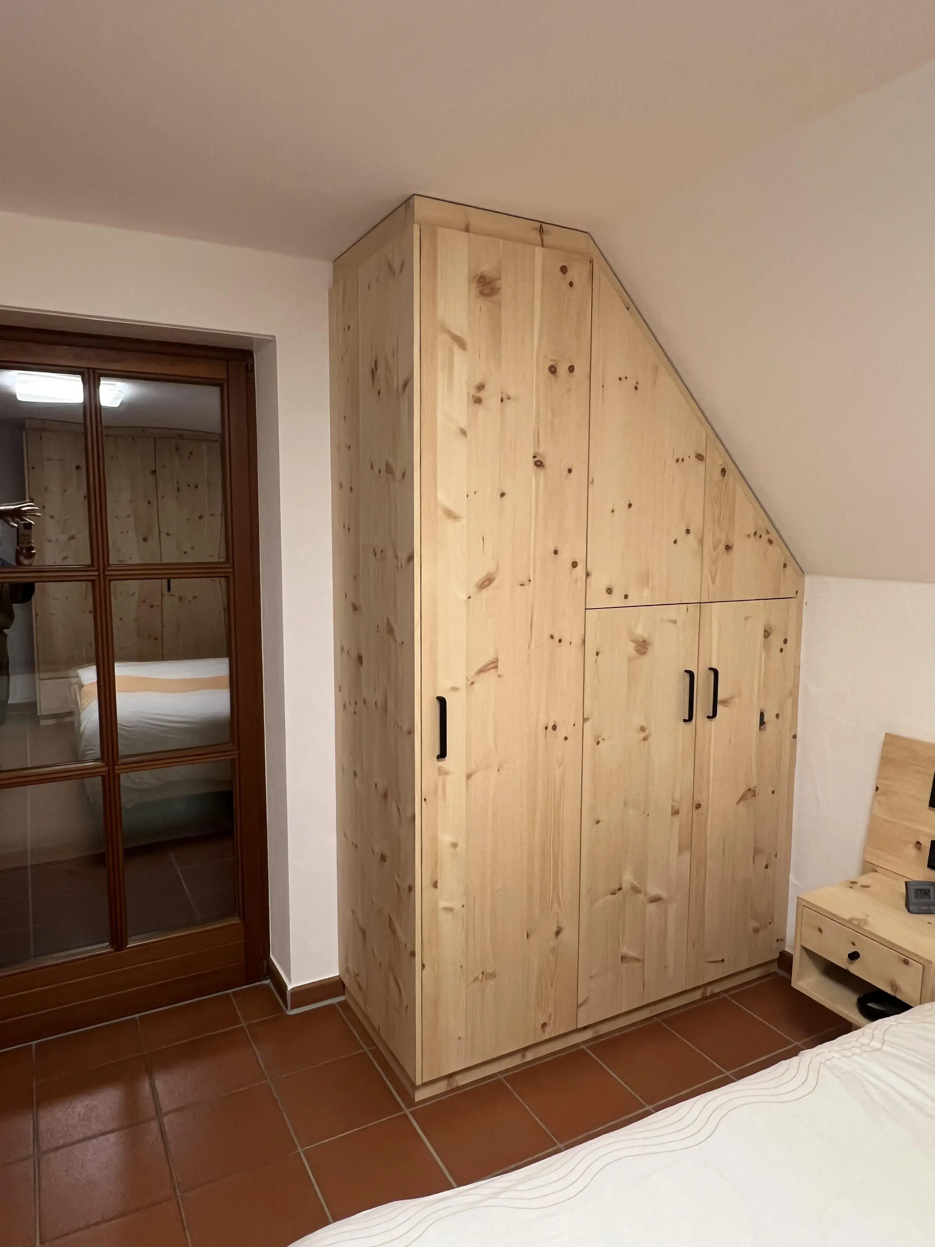 Schrank aus hellem Holz unter Dachschräge in einem Schlafzimmer mit Fliesenboden.