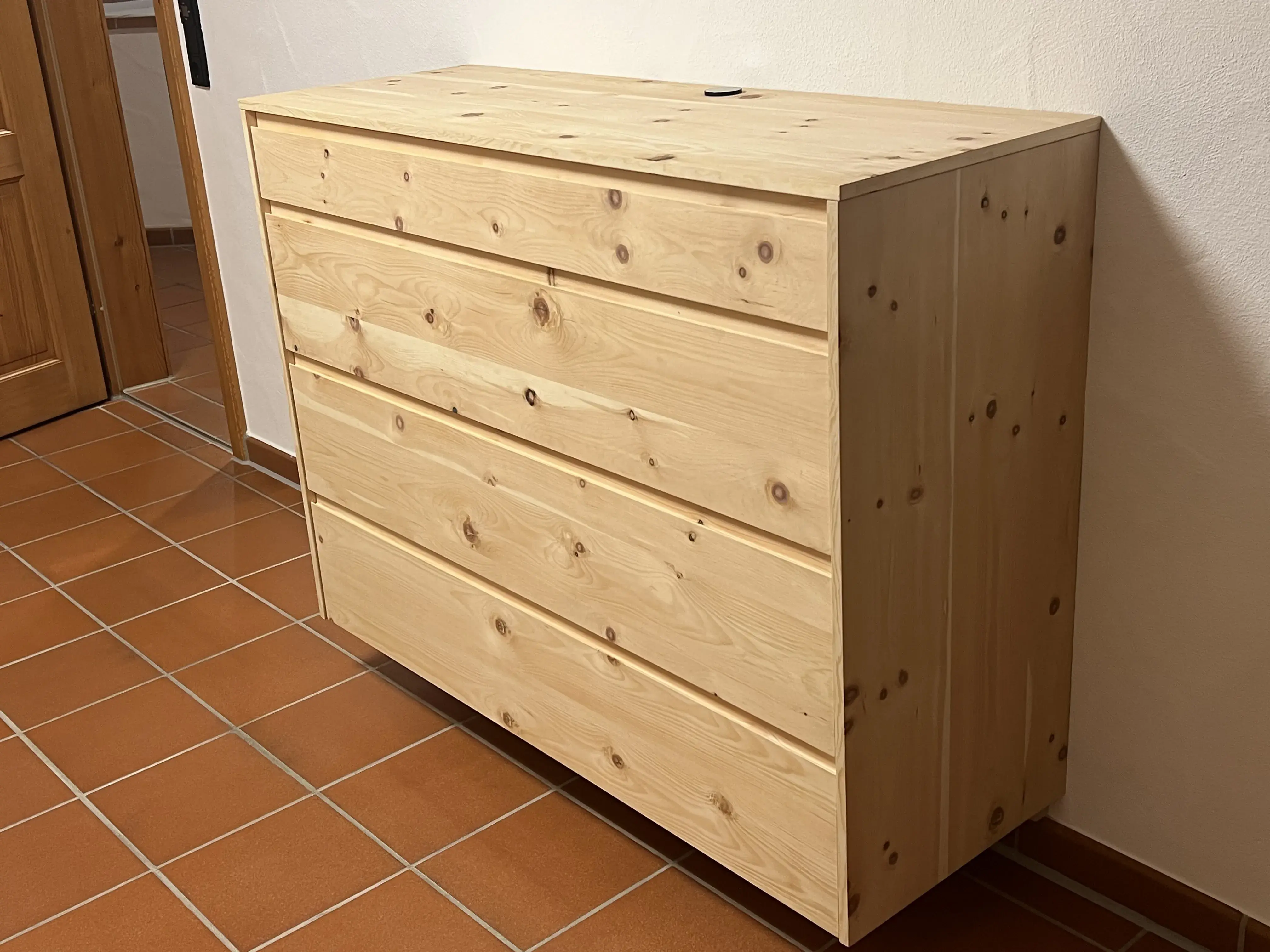 Helle Holzkommode mit vier Schubladen auf braunen Fliesenboden.