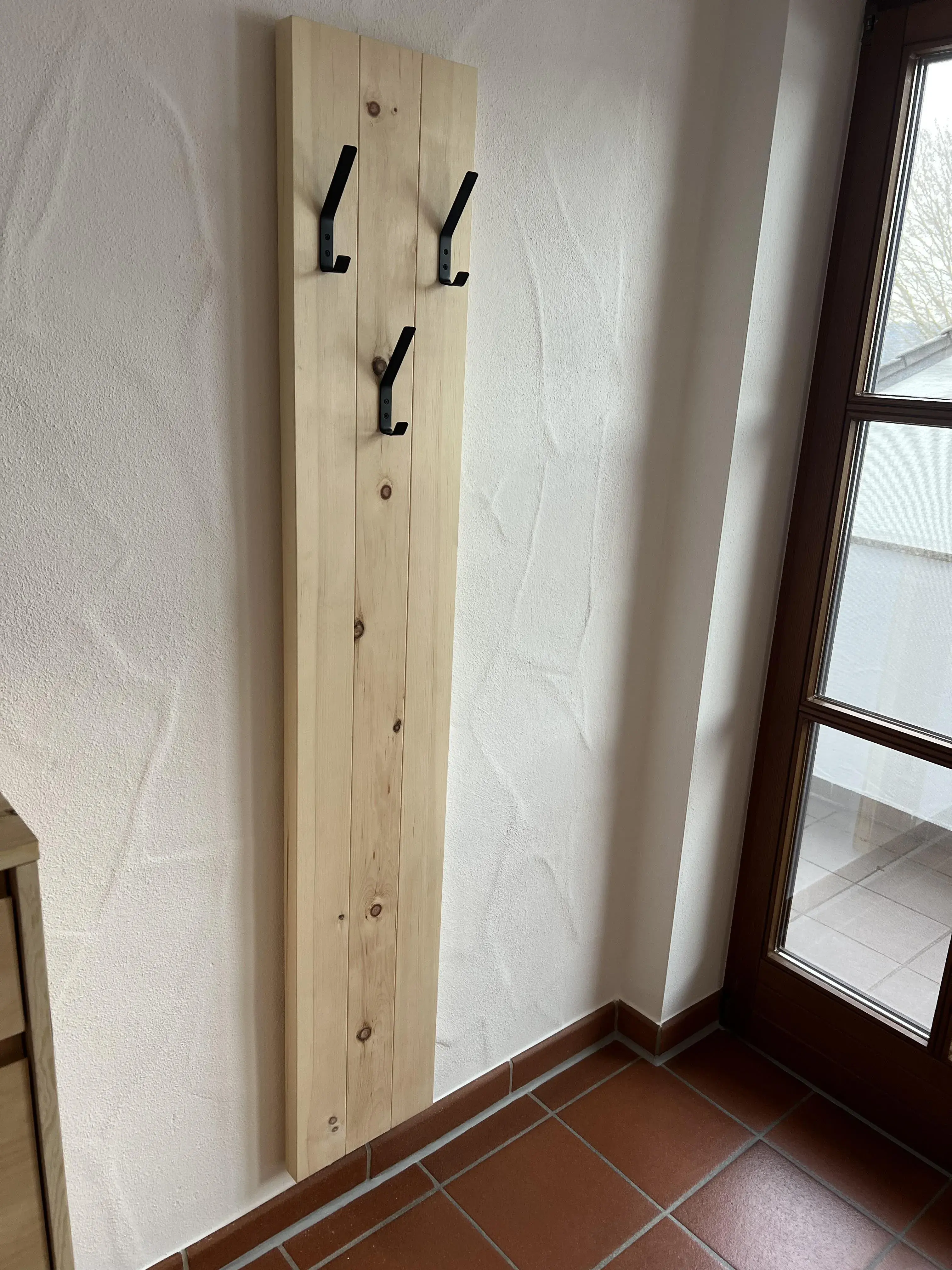 Schmaler Garderobenständer aus Holz mit vier schwarzen Haken an einer Wand im Flur.
