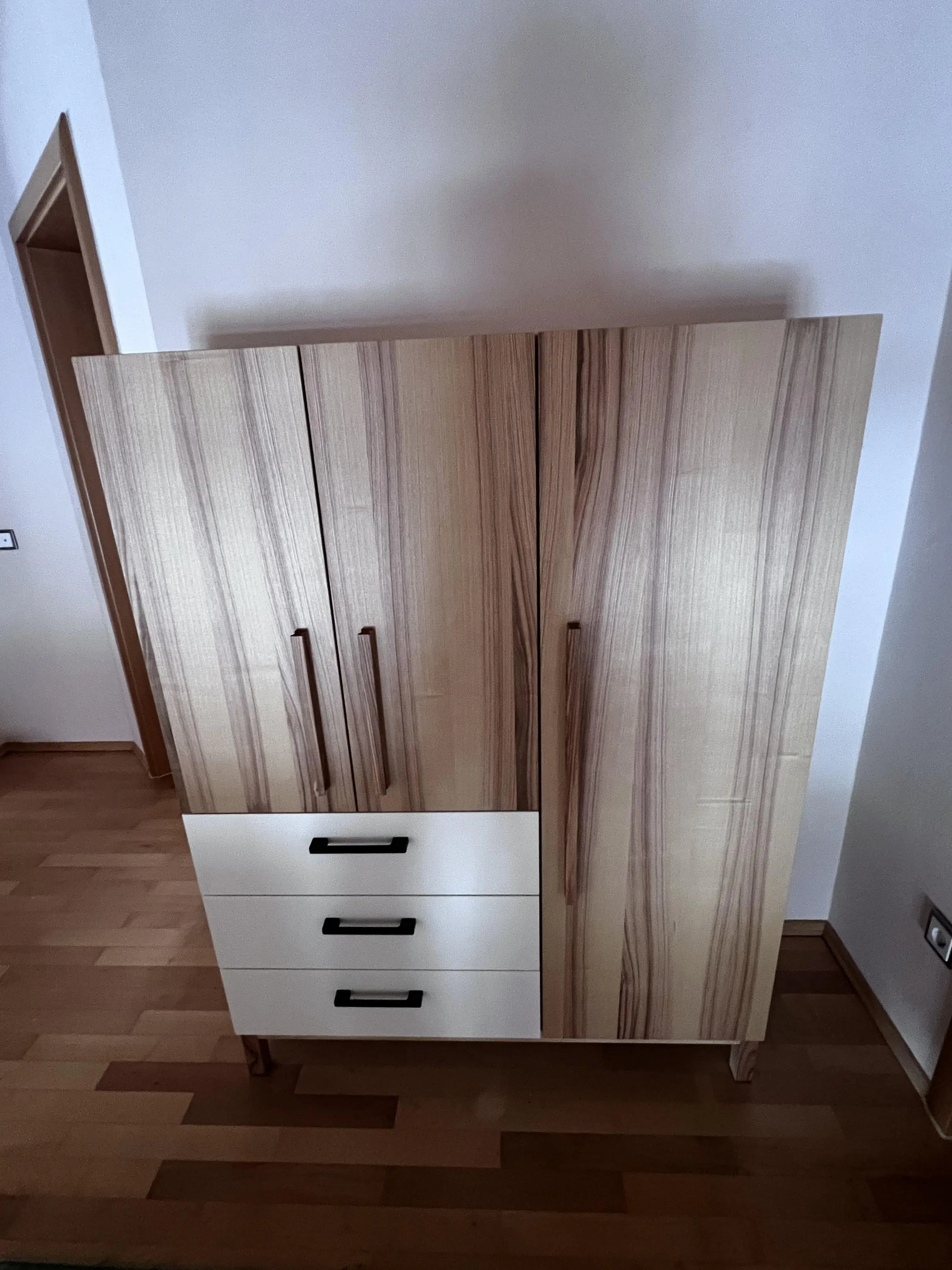 Holzkleiderschrank mit drei weißen Schubladen und zwei Türen, steht auf Parkettboden.