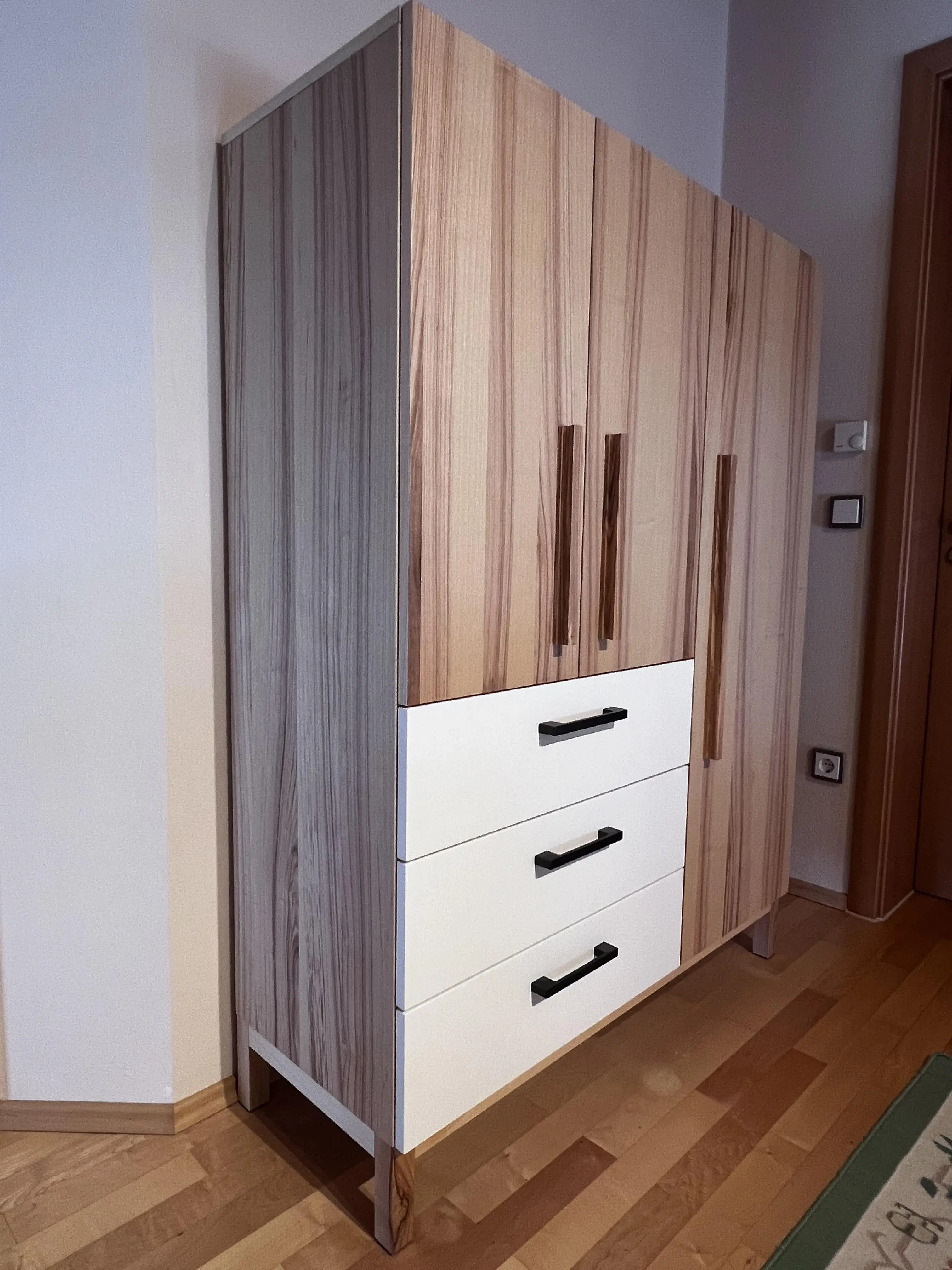 Holzschrank mit drei weißen Schubladen und zwei Türen, steht auf Holzboden.