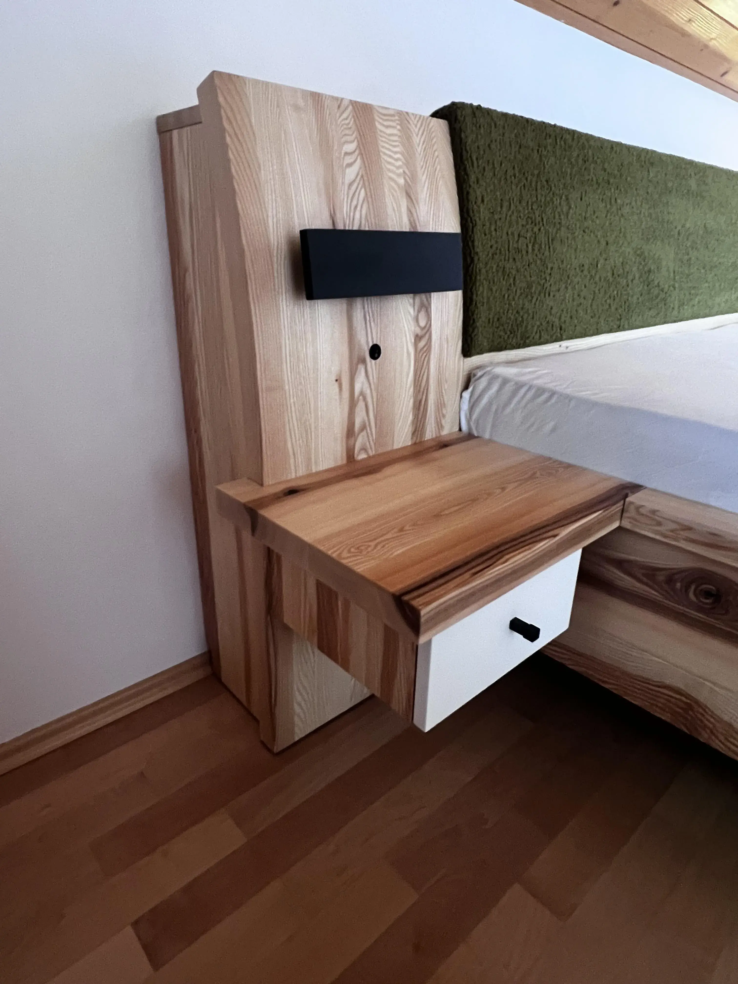 Nachttisch aus Holz mit Schublade, daneben Bett und grünem Kopfteil.