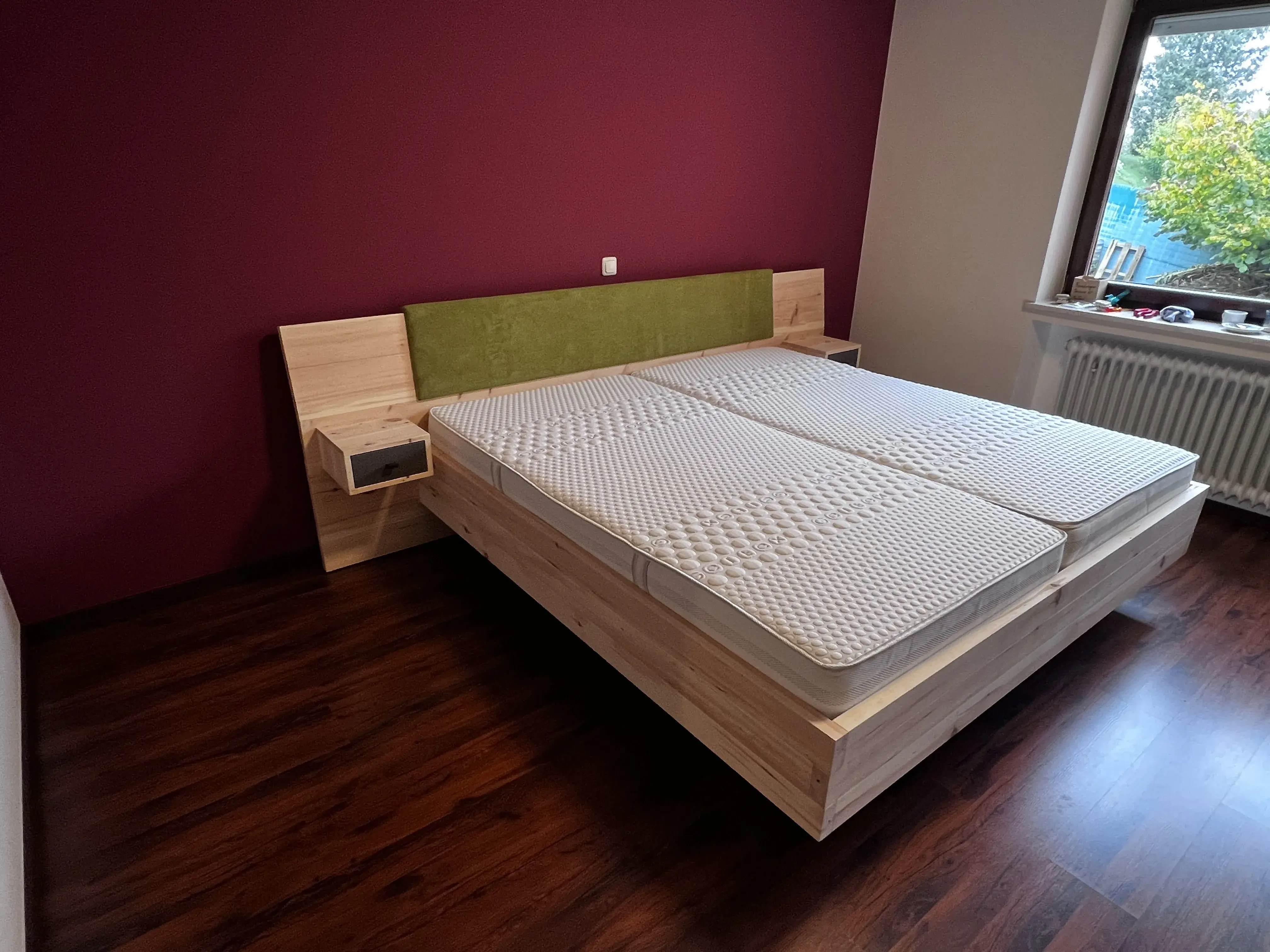 Modernes Doppelbett aus Holz mit grünem Polsterkopfteil vor bordeauxfarbener Wand.