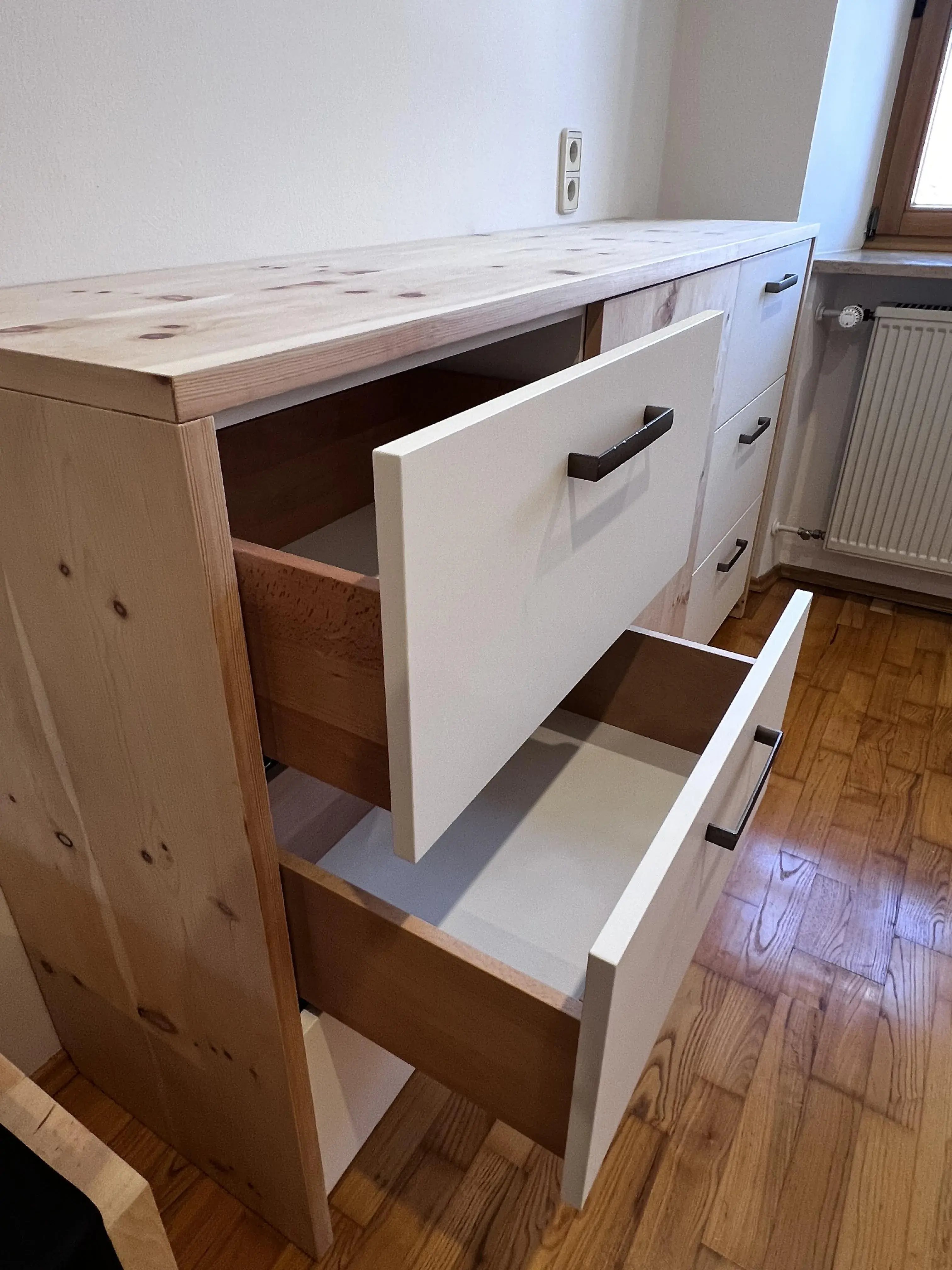 Holzkommode mit zwei offenen weißen Schubladen auf Parkettboden neben einem Fenster.