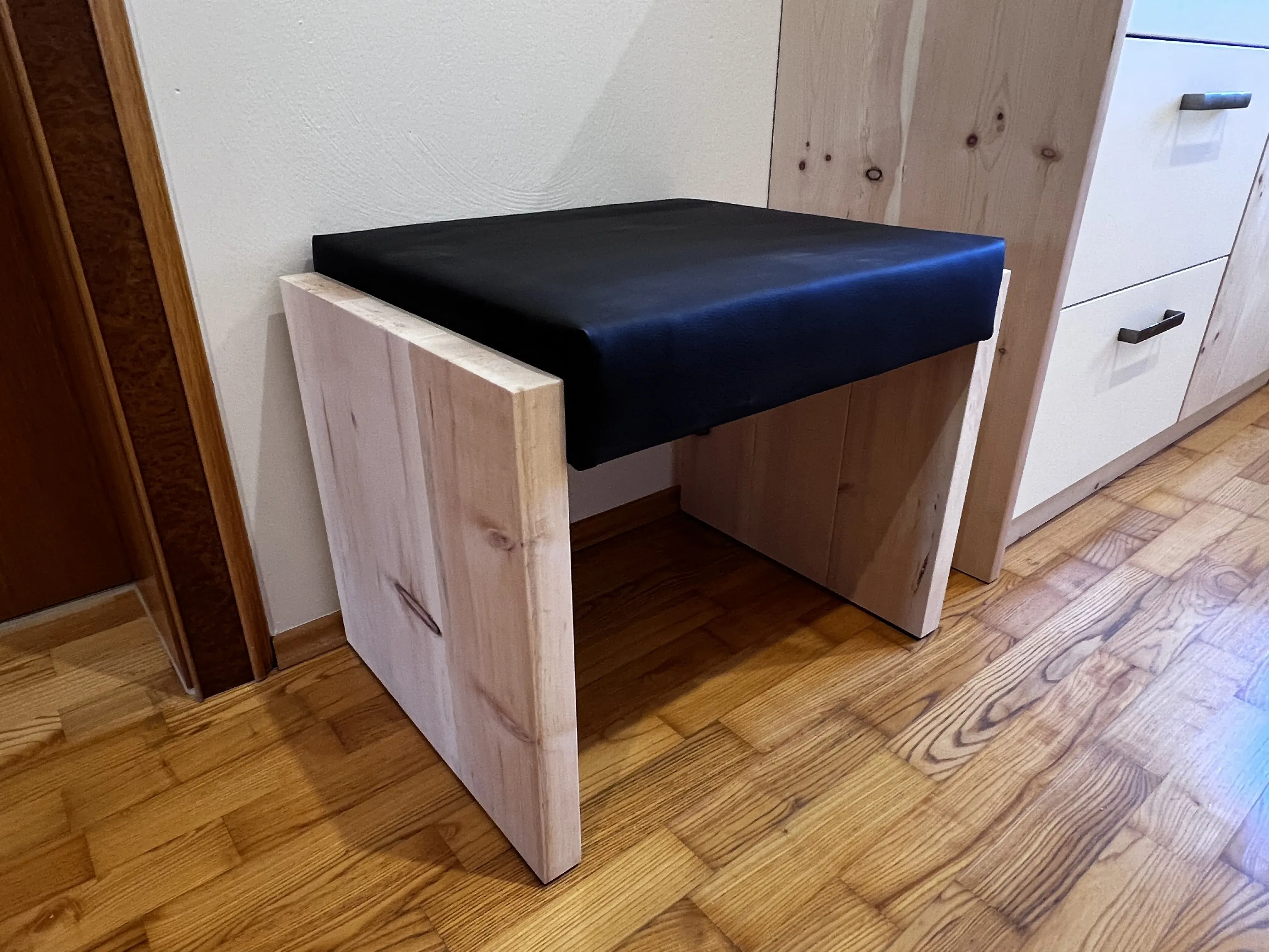 Holzhocker mit schwarzem Sitzpolster auf Parkettboden neben einem Schrank