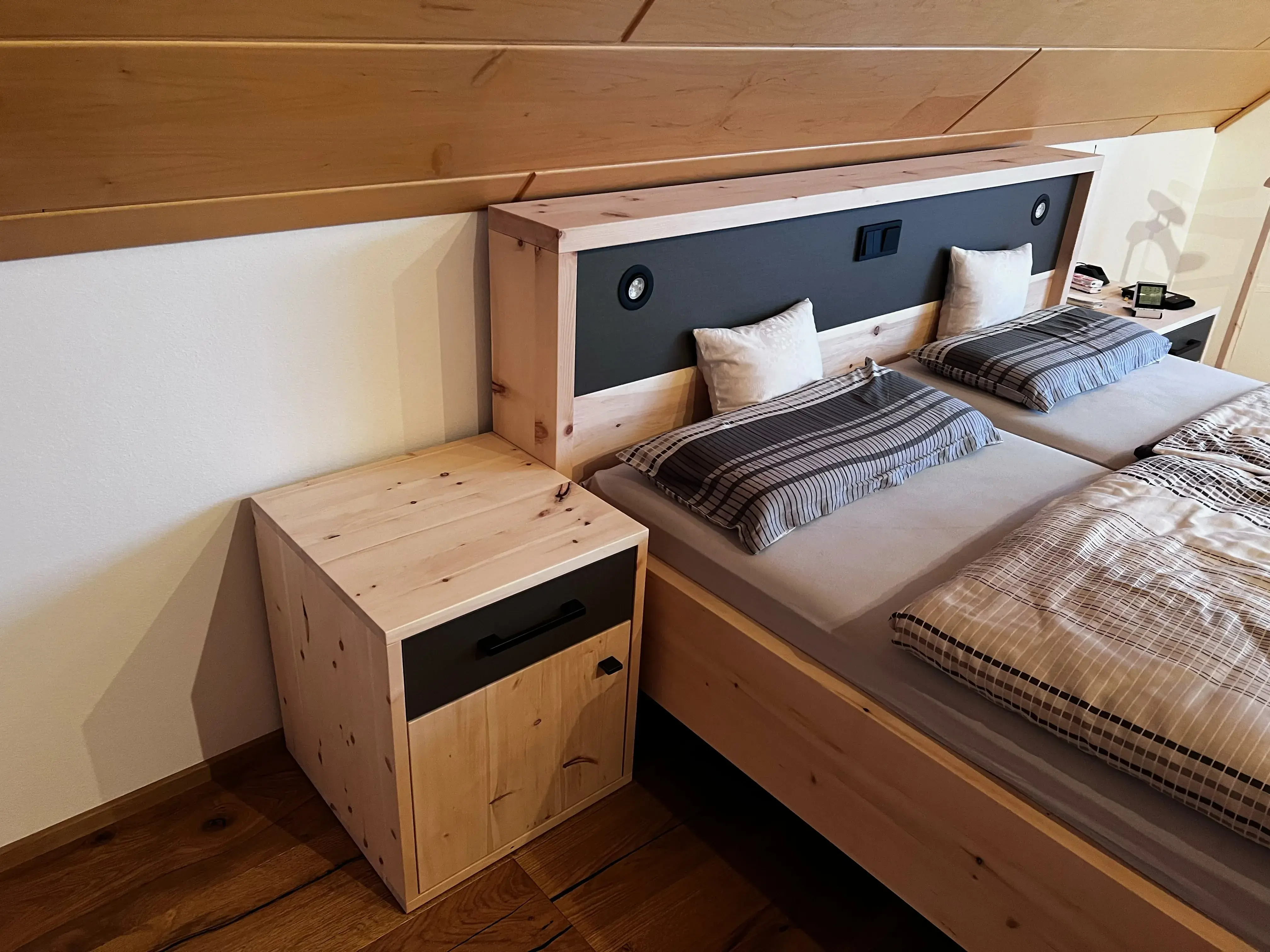 Holzbett mit Nachttisch, zwei Kissen und gestreifter Bettwäsche in einem Dachzimmer.