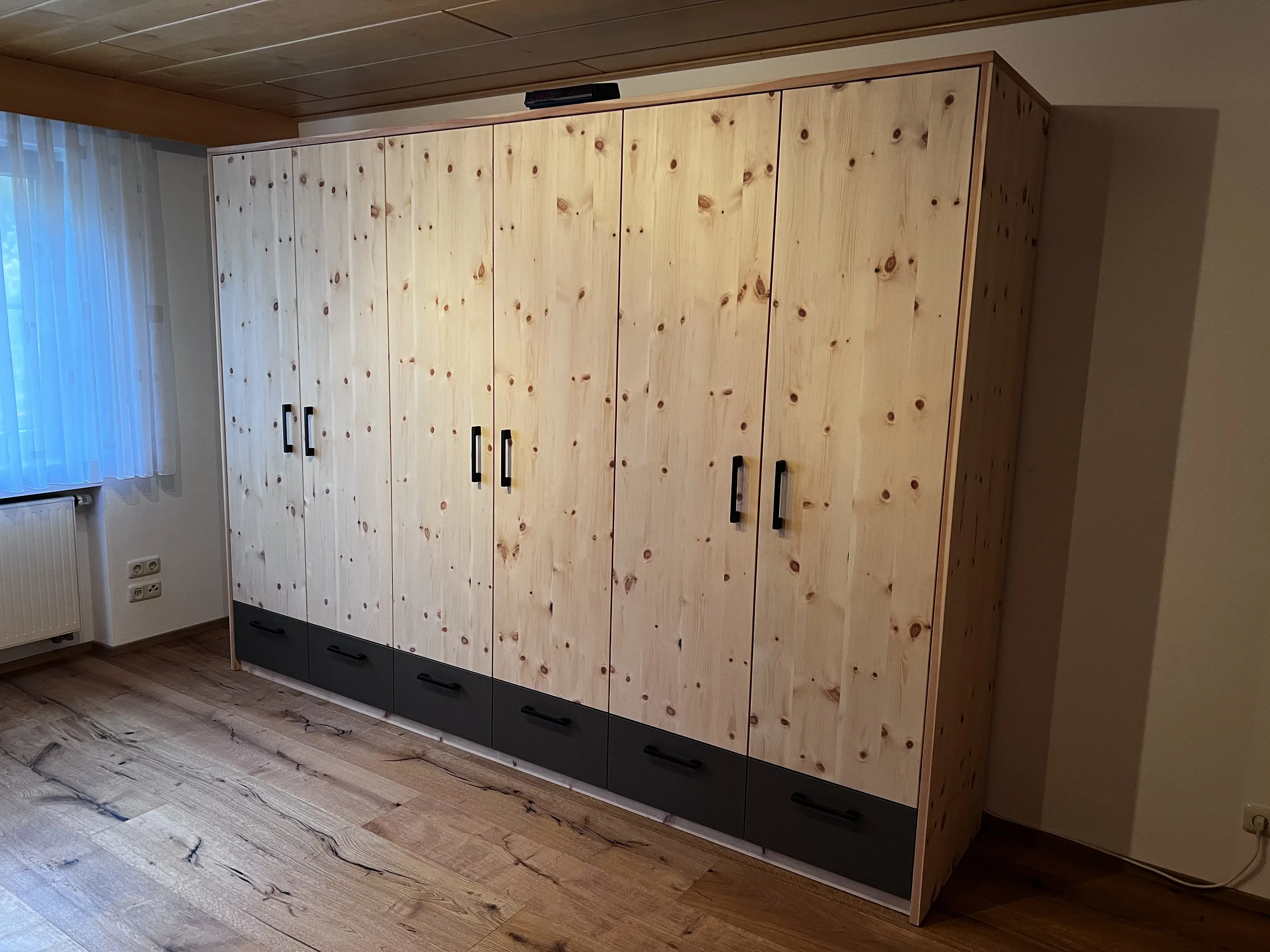 Großer Kleiderschrank aus hellem Holz mit schwarzen Griffen in einem Zimmer mit Holzboden.