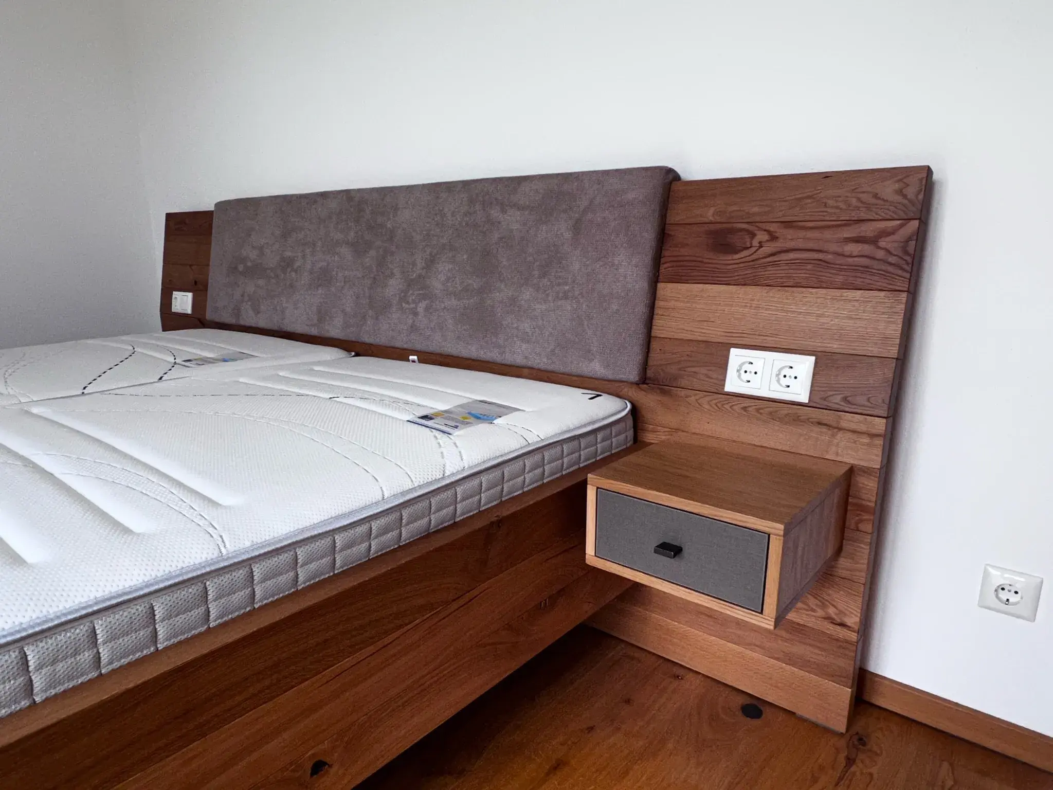 Holzbett mit grauem Polsterkopfteil und integriertem Nachttisch mit Schublade.