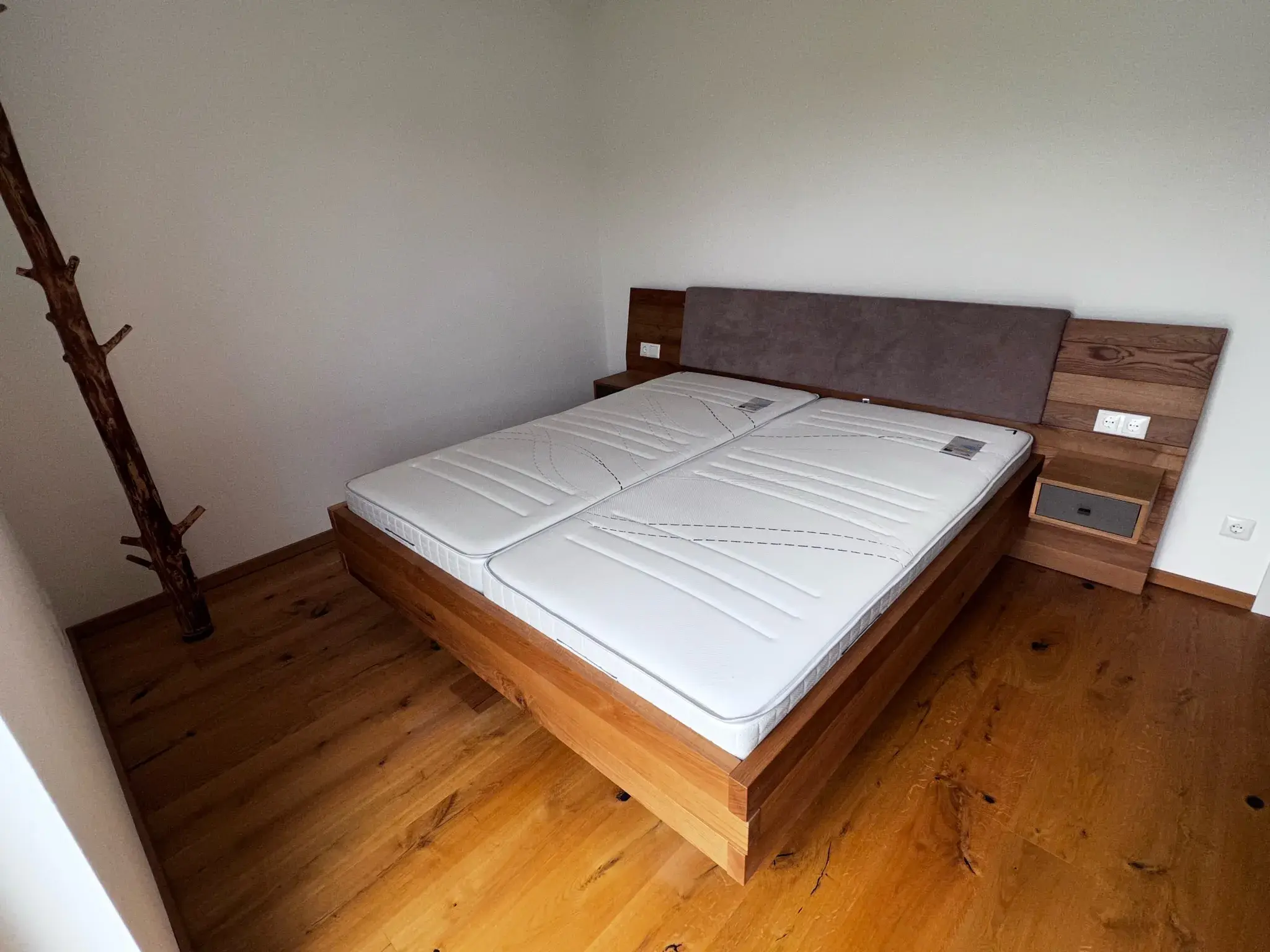 Doppelbett mit Holzrahmen und weißen Matratzen in einem schlichten Schlafzimmer.