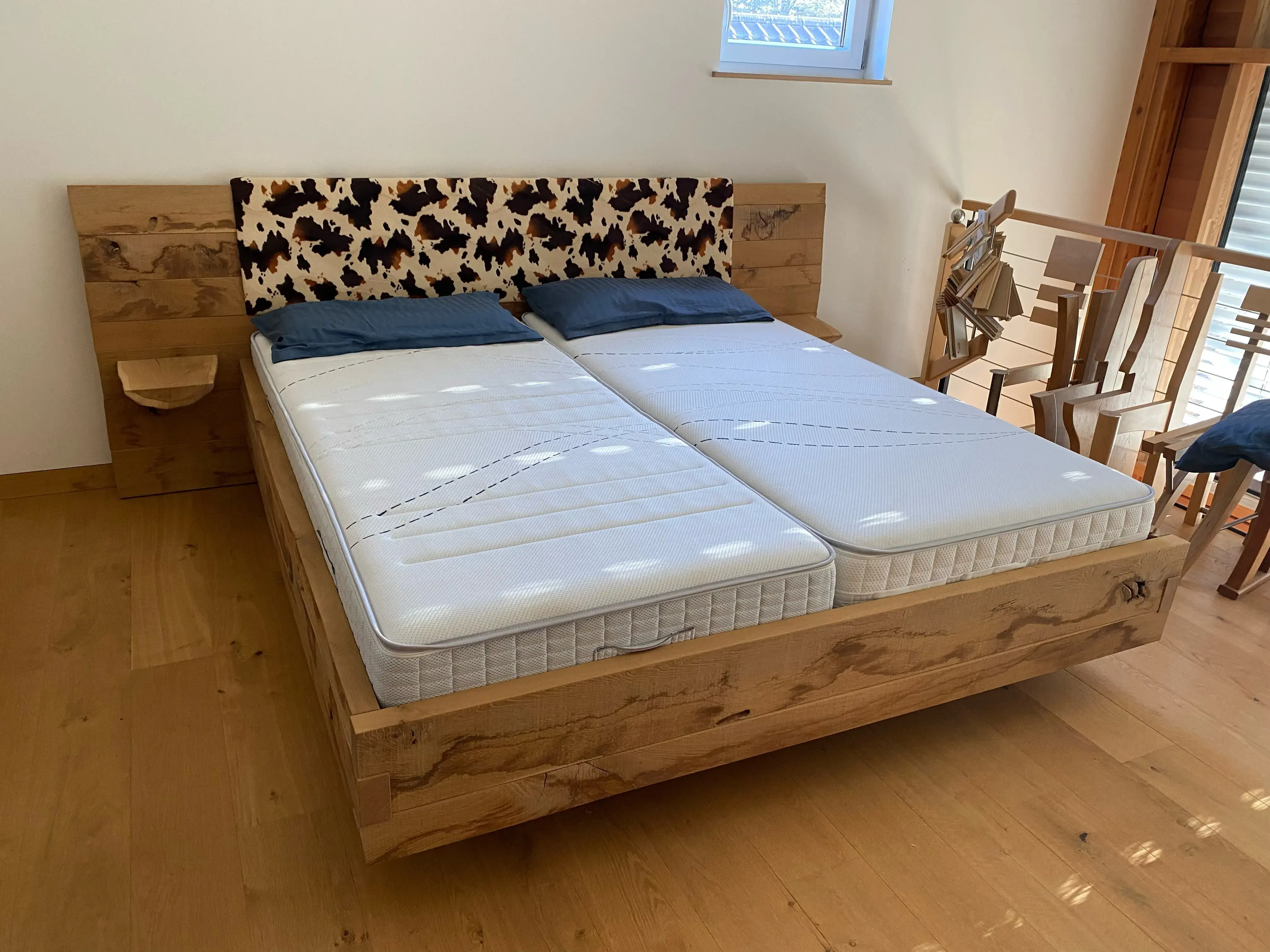 Doppelbett aus Holz mit Kuhfell-Kopfteil und zwei Matratzen, Holzfußboden, Tageslicht.