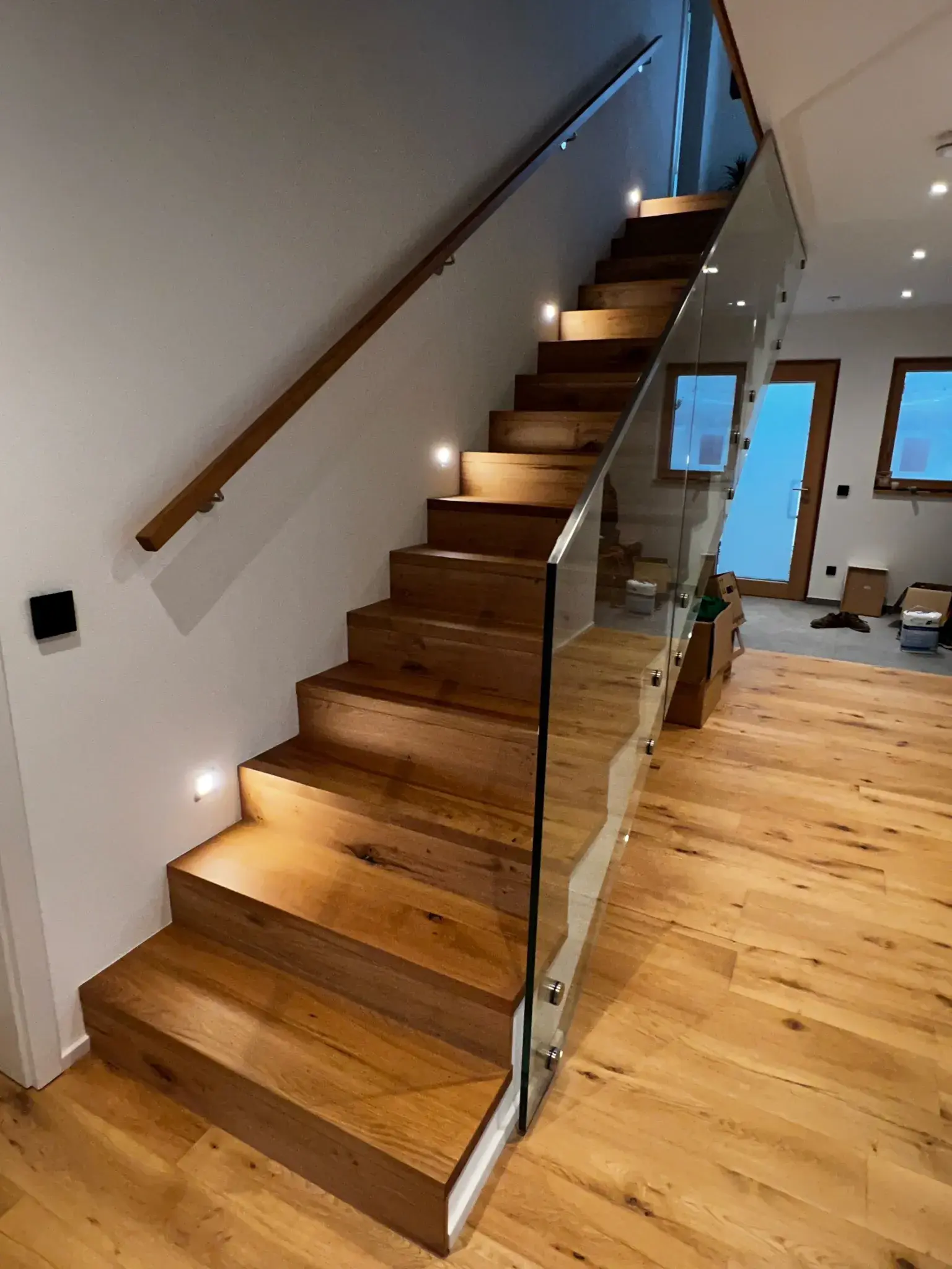 Moderne Holztreppe mit Glasgeländer und integrierter Stufenbeleuchtung in einem Hausflur.