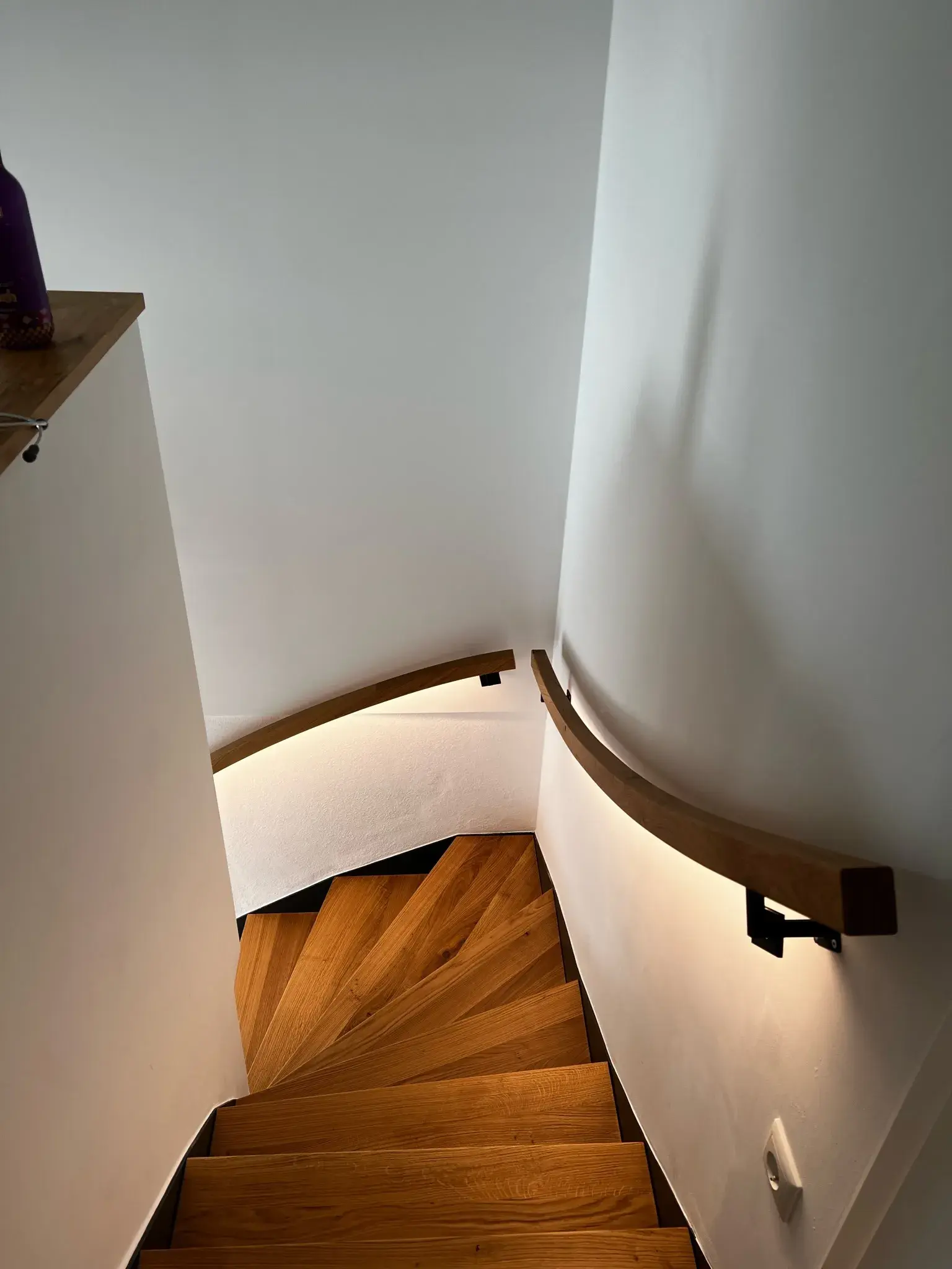 Holztreppe mit hellem Handlauf in einem engen, weiß gestrichenen Treppenhaus.