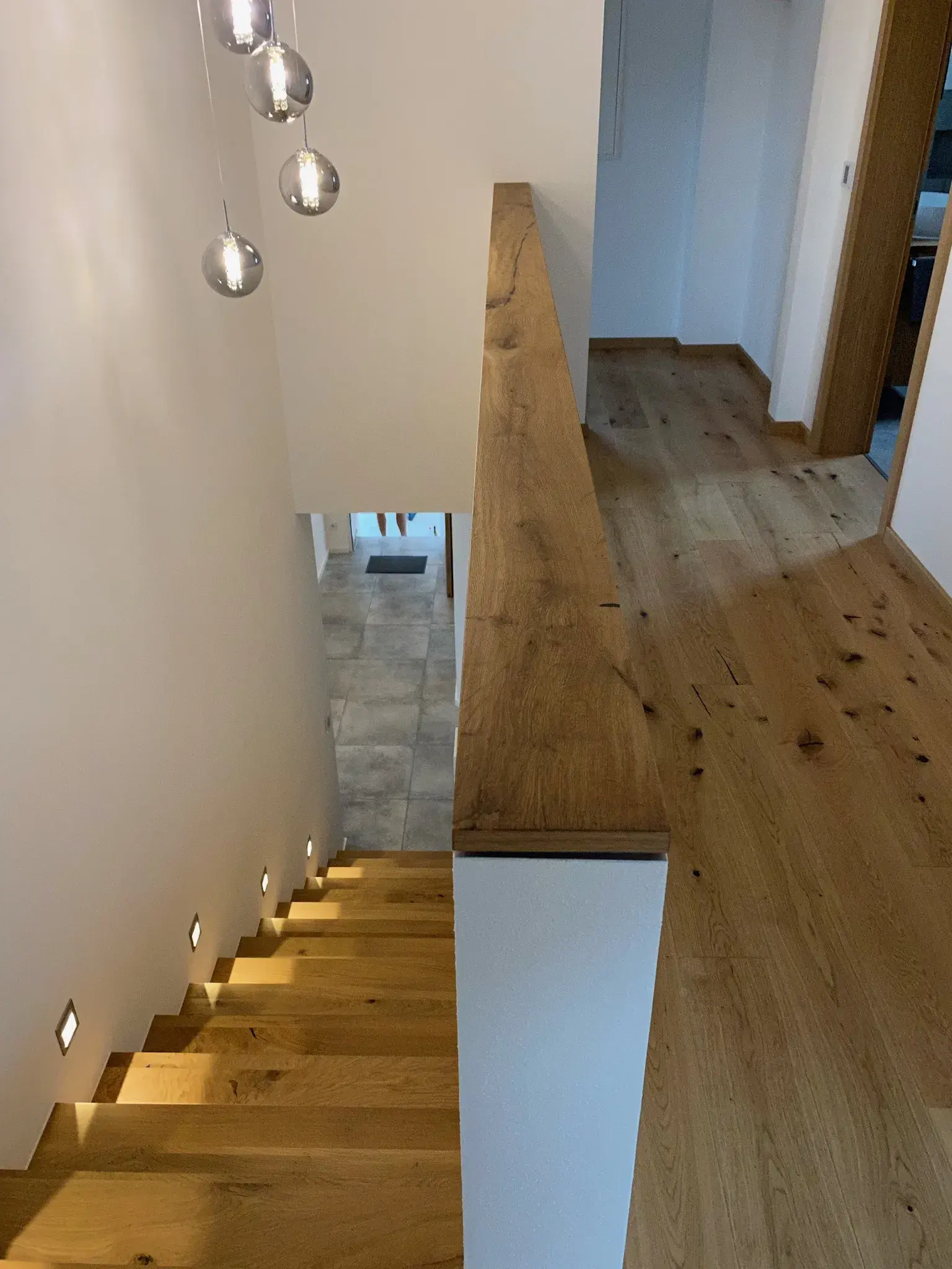 Holztreppe mit seitlichen Wandlampen, oben Flur mit Parkett, moderne Pendelleuchten.