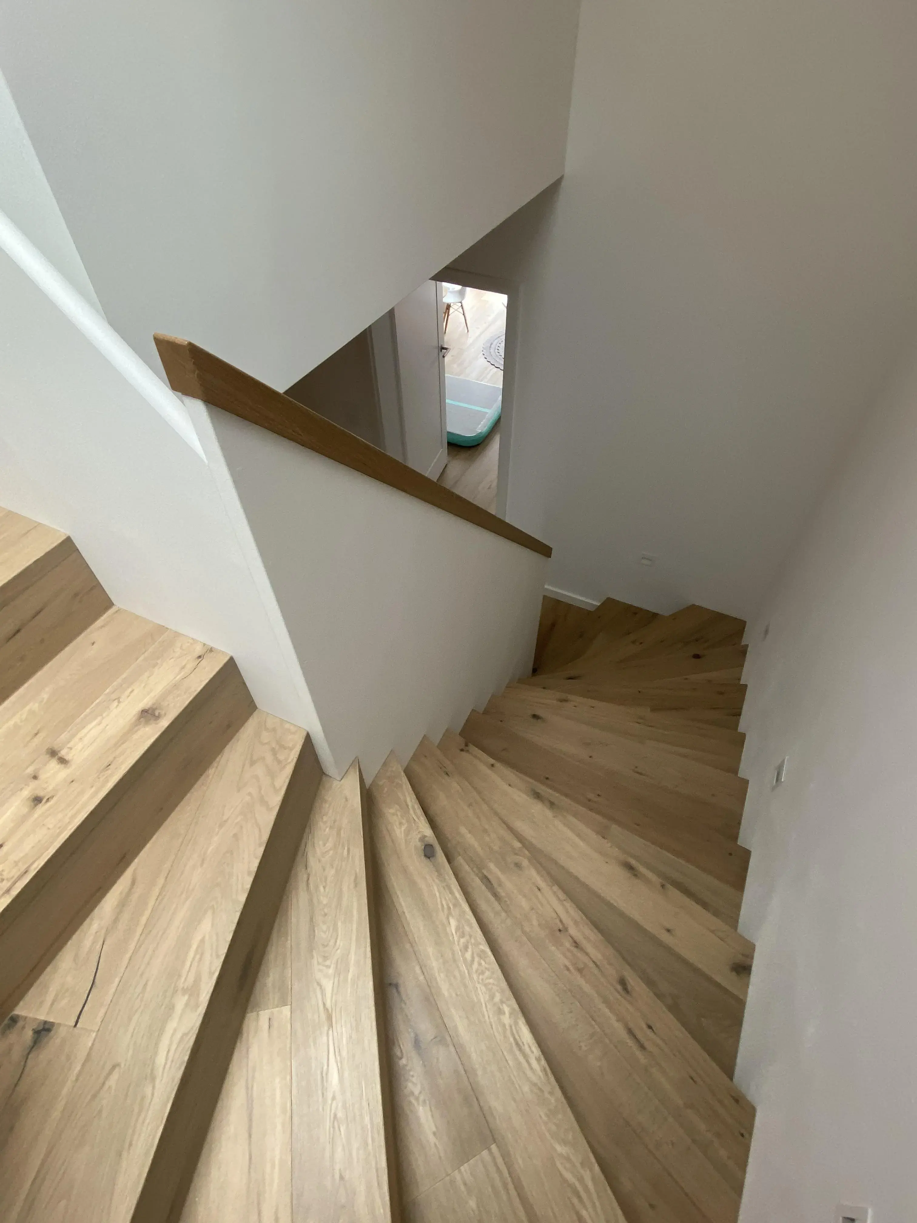 Holztreppe mit weißer Wand, Blick nach unten, Tageslicht durch Türöffnung.