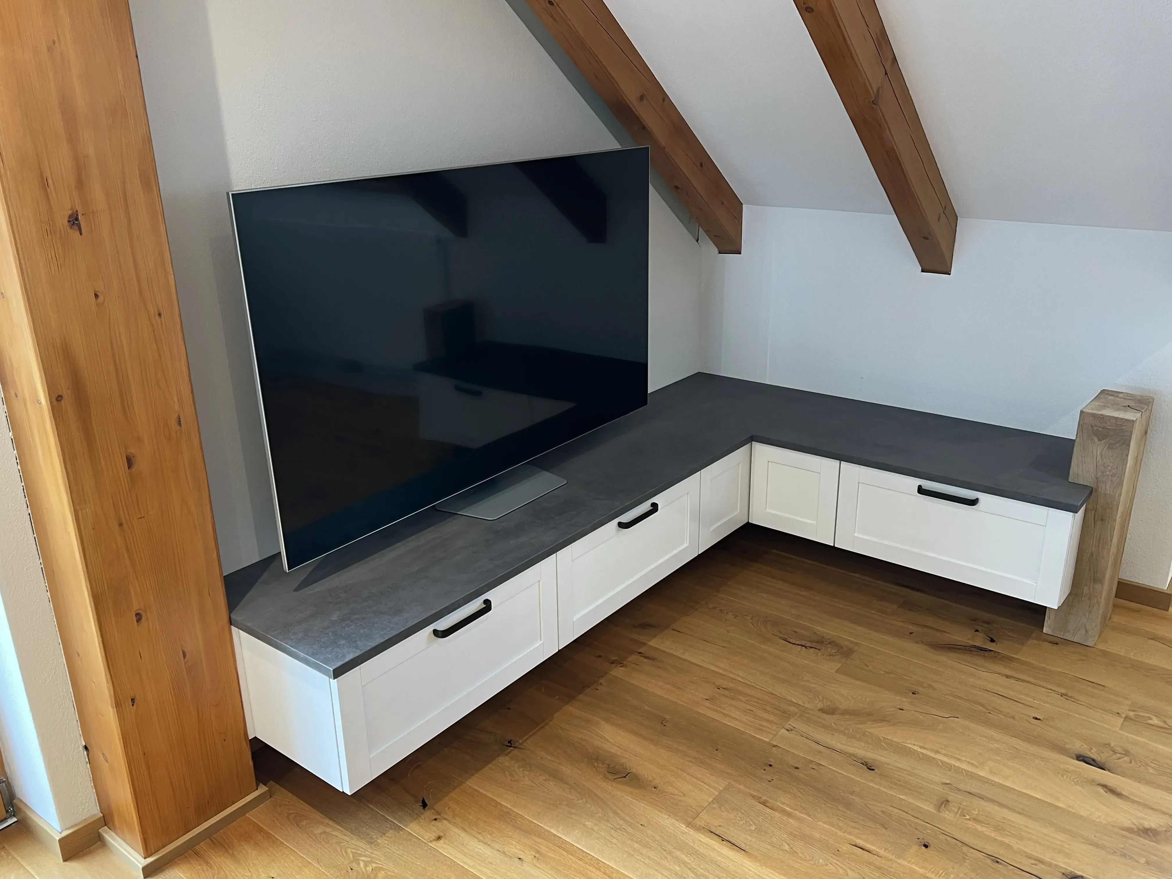 Großer Fernseher auf einem modernen Eck-TV-Schrank unter Dachschrägen, Holzboden.